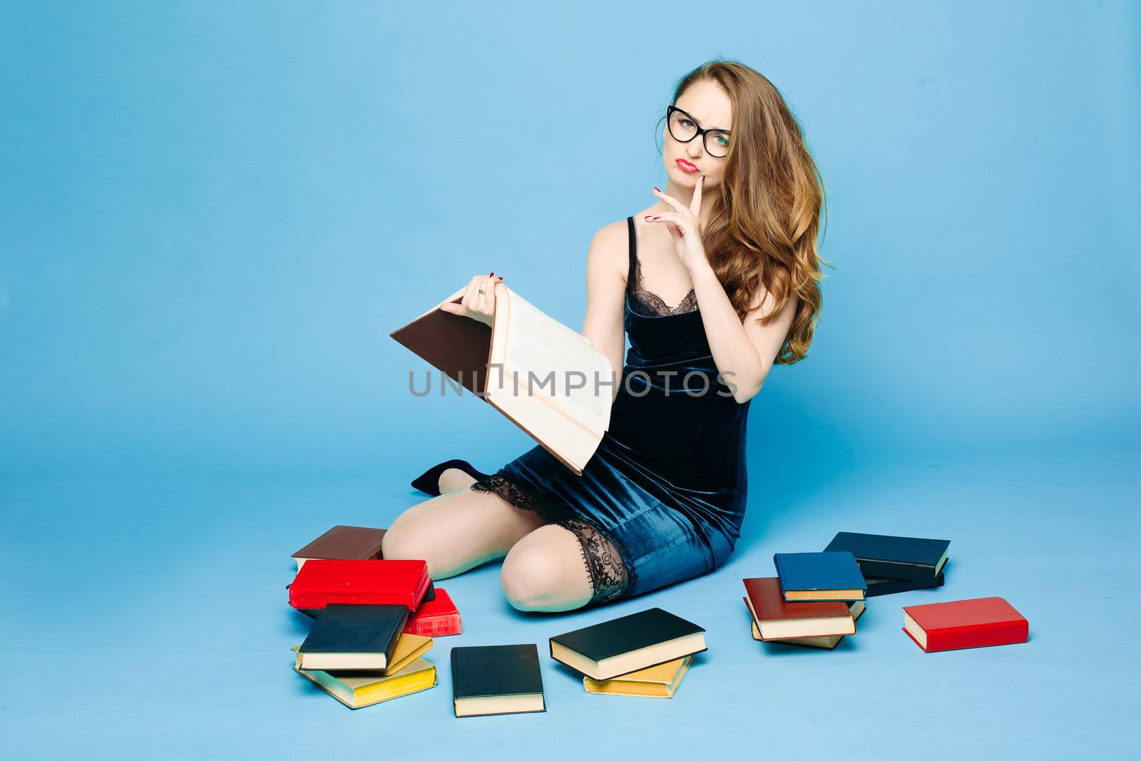 Sexy and beautiful female teacher in dress and eyeglasses, sitting on floor, posing and reading book, gesturing by hands, surprised looking up. Stylish pretty teacher or seductive female student.