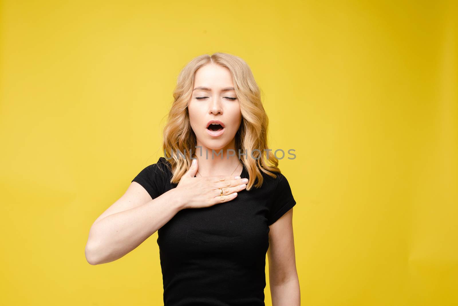 Stock photo of a blonde Caucasian woman in black t-shirt holding hand on chest demonstrating problem with breathing. Asthma attack, respiratory problem, sore throat. Isolate on yellow.