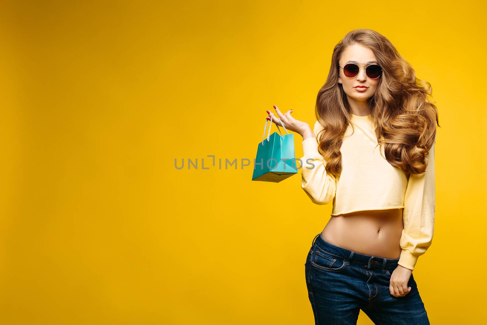 Fashionable sexy brunette woman with volumed wavy hair holding little blue paper bag with present, after shopping and posing at camera. Stylish woman in sunglasses and yellow crop sweatshirt.
