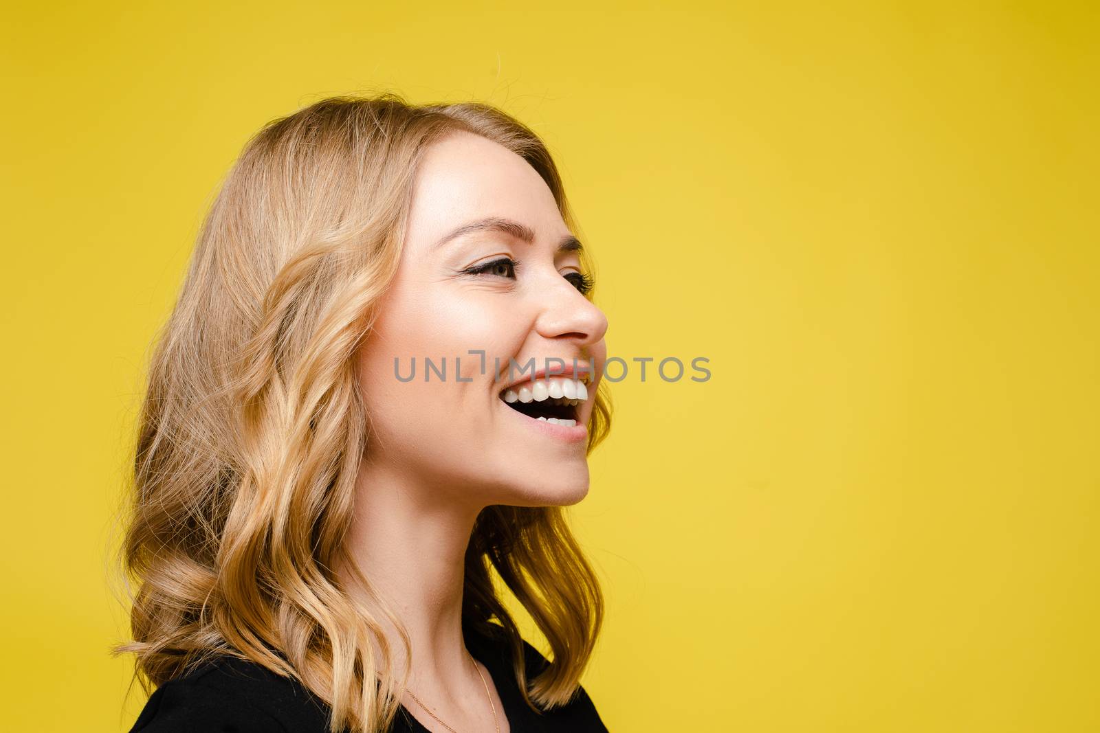 Beautiful caucasian woman with light wavy hair in a black t-shirt laughs isolated on yellow background