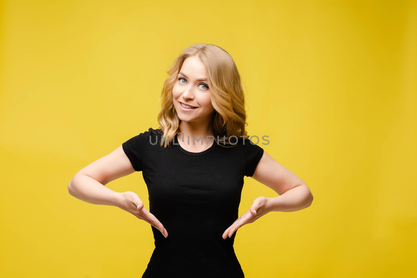Cheerful young female with light wavy hair in a black t-shirt, blue pants smiles isolated on yellow background