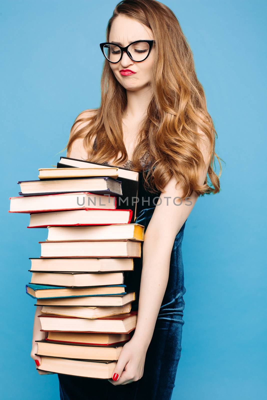 Portrait of emotionally shocked sexy teacher in dress with lace, eyeglasses, holding many books and surprised looking at camera. Young teacher or student screaming. Blue studio background.