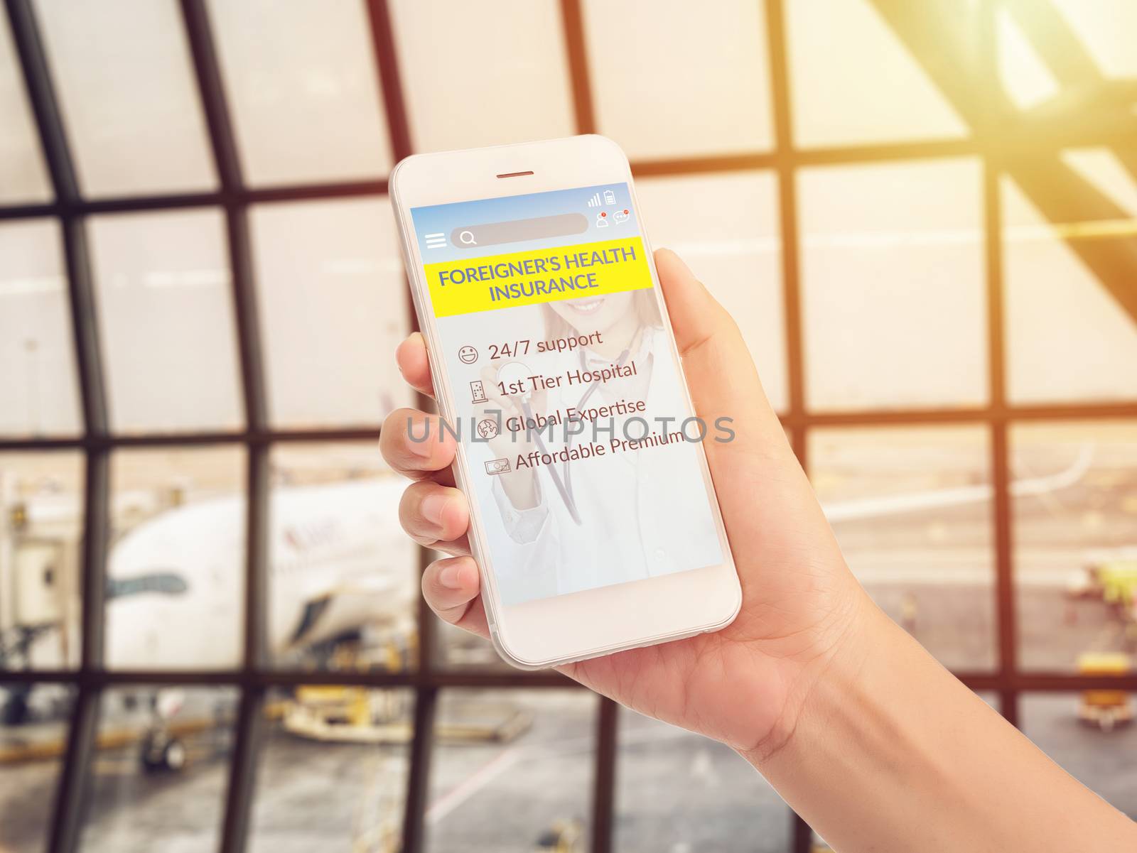 hand holding mobile phone with advertising screen for foreigner's health insurance application with good service and covered benefit condition, airport terminal with airplane parking at background