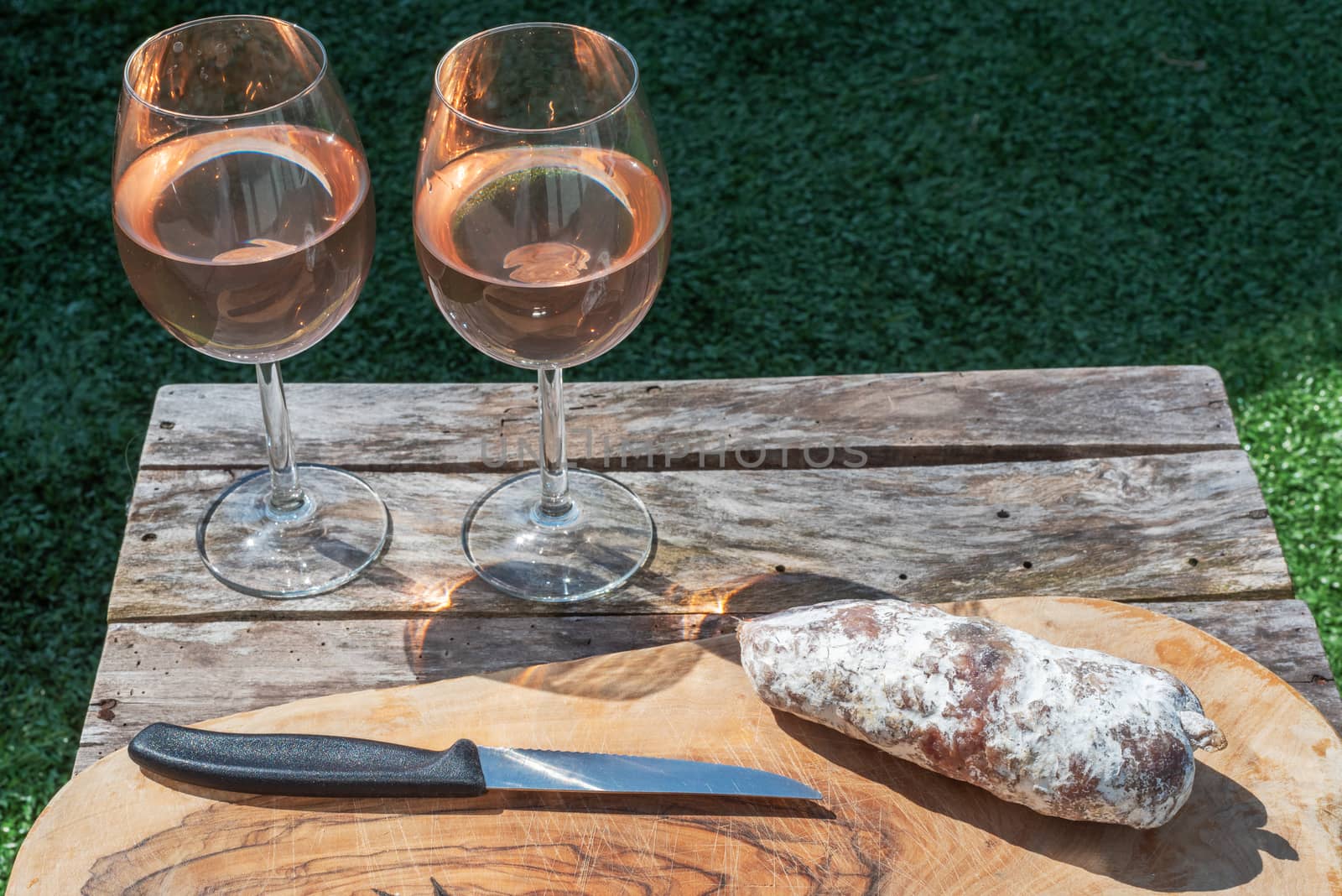 Two glasses of rose wine and a dried sausage with a knife on a wooden table in a garden on a sunny day