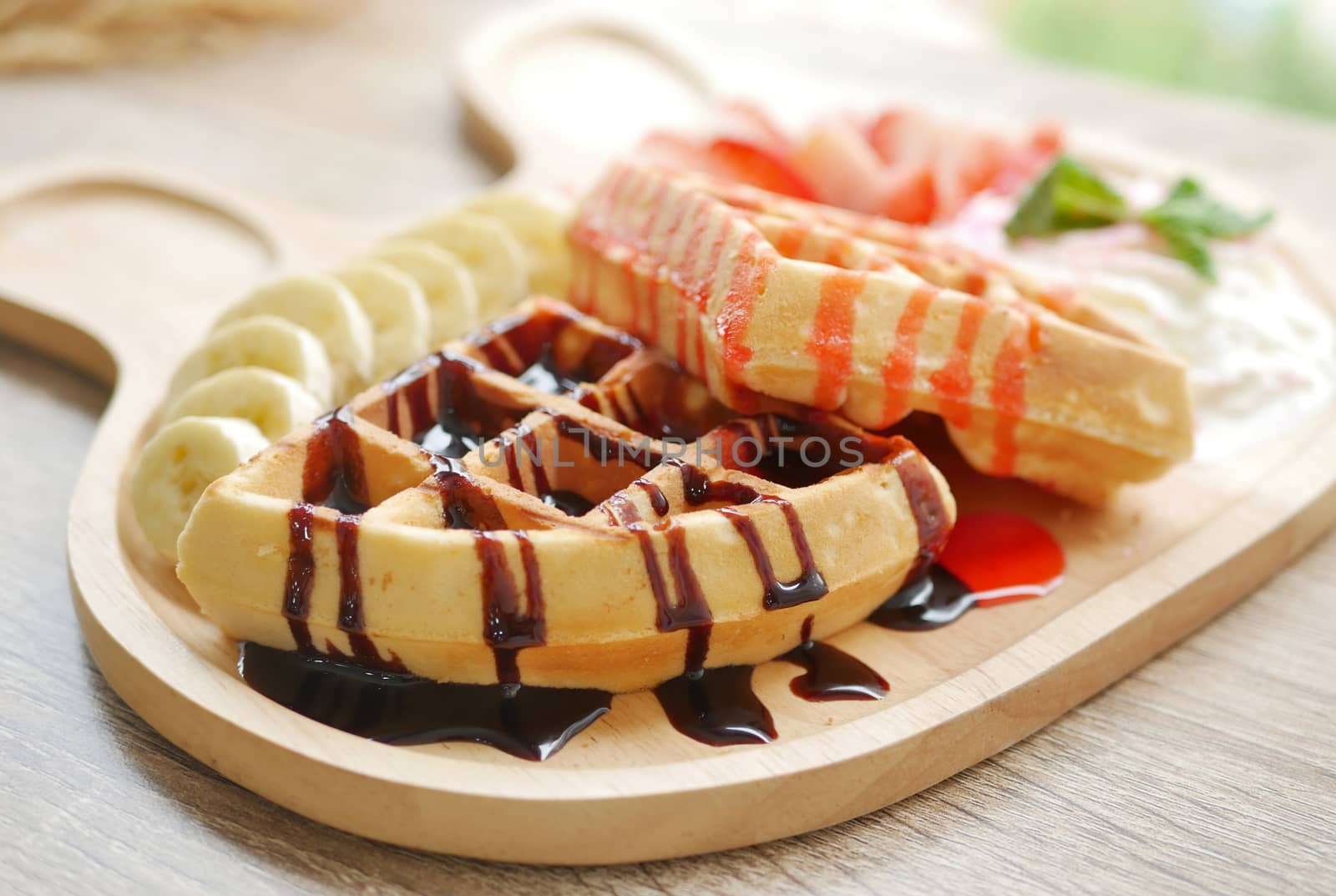 tasty waffle topped with strawberry syrup and chocolate sauce, side dishes are fresh strawberry chopped , banana , whipped cream and ice cream served on cute wooden plate