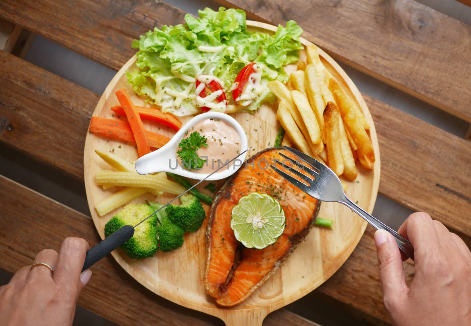 salmon steak served on wooden plate ,side dishes are boiled vegetables , French fries , fresh salad ,cream sauce and piece of lemon on top by asiandelight
