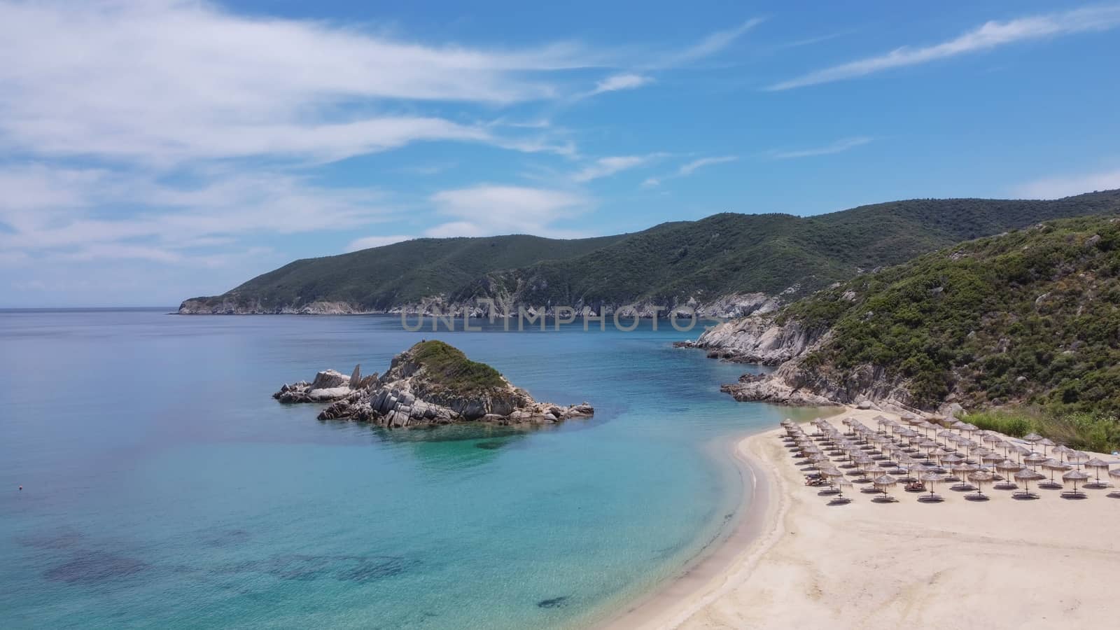 Aerial view of Sithonia Chalkidiki peninsula with green plantation by crystal clear waters, sea beds with umbrellas & small rocky island.