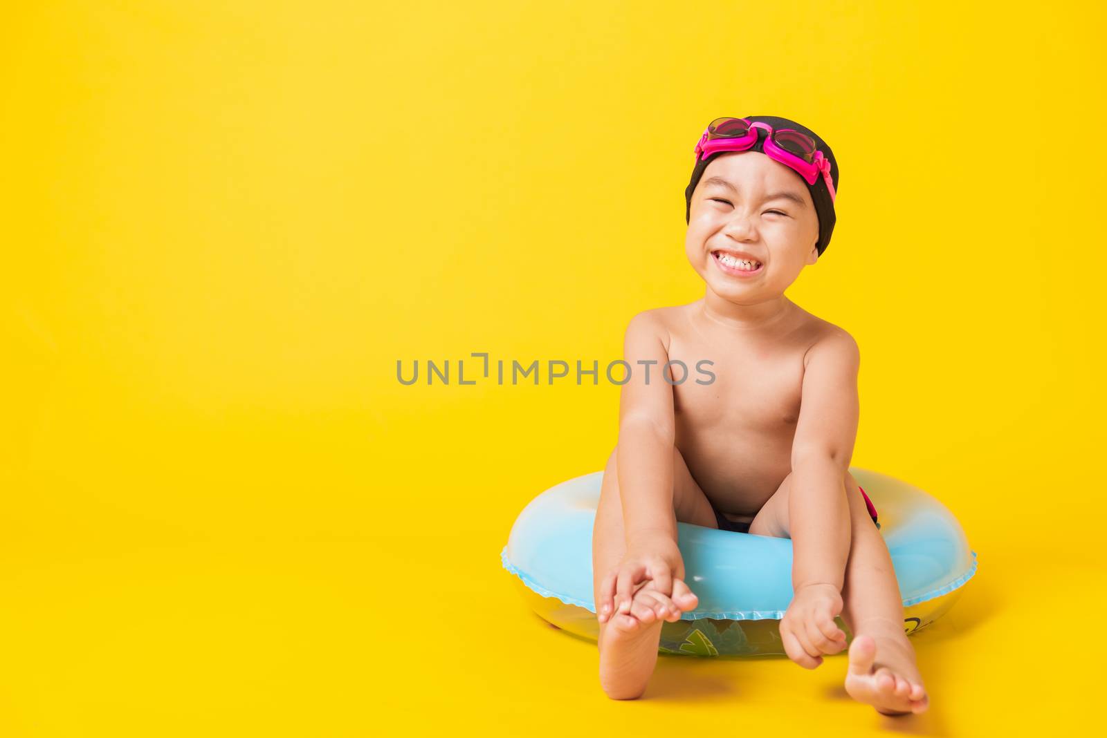 Summer vacation concept, Portrait Asian happy cute little child boy wear goggles, swimsuit hold beach blue inflatable ring, Kid have fun sit in inflatable, studio shot isolated yellow background