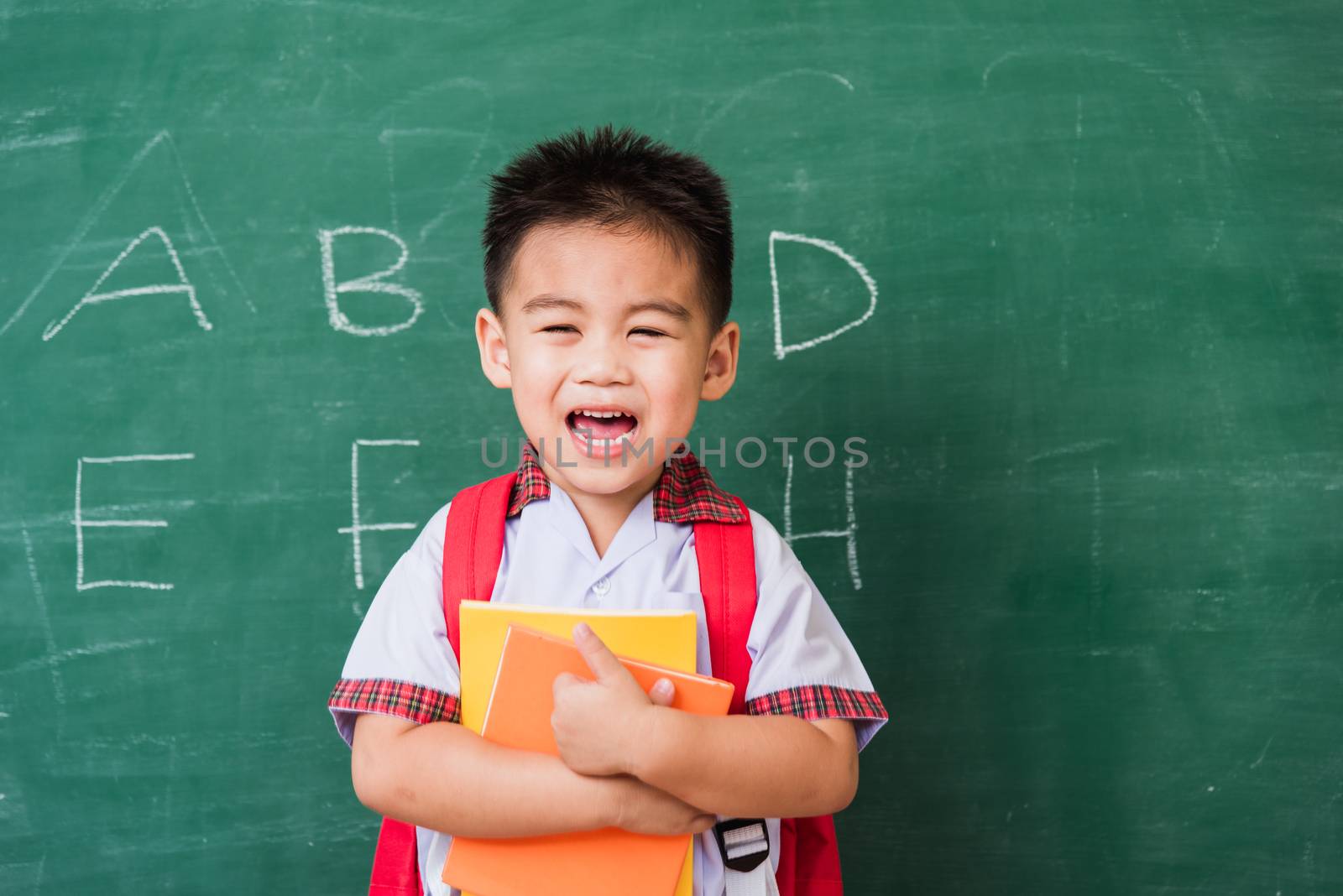 Back to School. Happy Asian funny cute little child boy kindergarten in student uniform with school bag and books smile show finger thumb up on green school blackboard, First time to school education