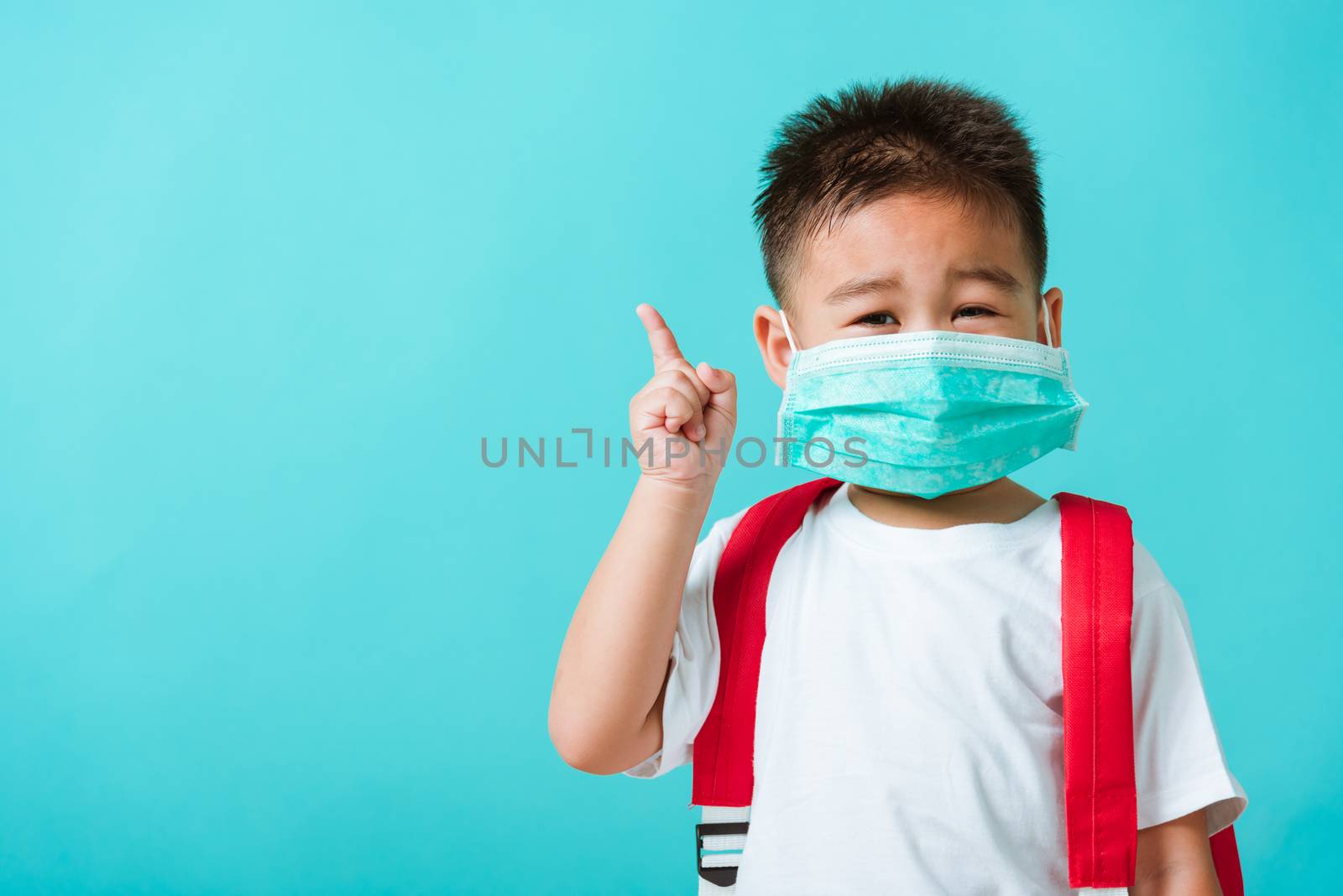 Back to school coronavirus Covid-19 education. Portrait Asian cute little child boy kindergarten wear face mask protective and school bag pointing finger to side, studio shot isolated blue background