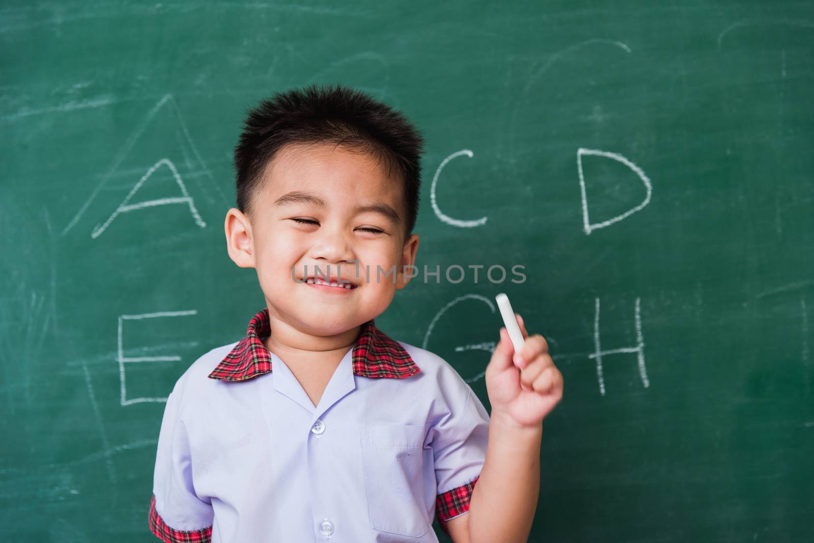 Back to School. Happy Asian funny cute little child boy kindergarten preschool smile in student uniform hold white chalk after write ABC with on green school blackboard, First time to school education