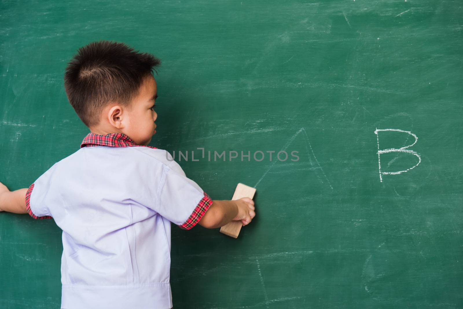 Back to School. Back of Asian cute little child boy kindergarten preschool in student uniform wiping clean or erase chalk on green school blackboard with sponge, First time to school education concept