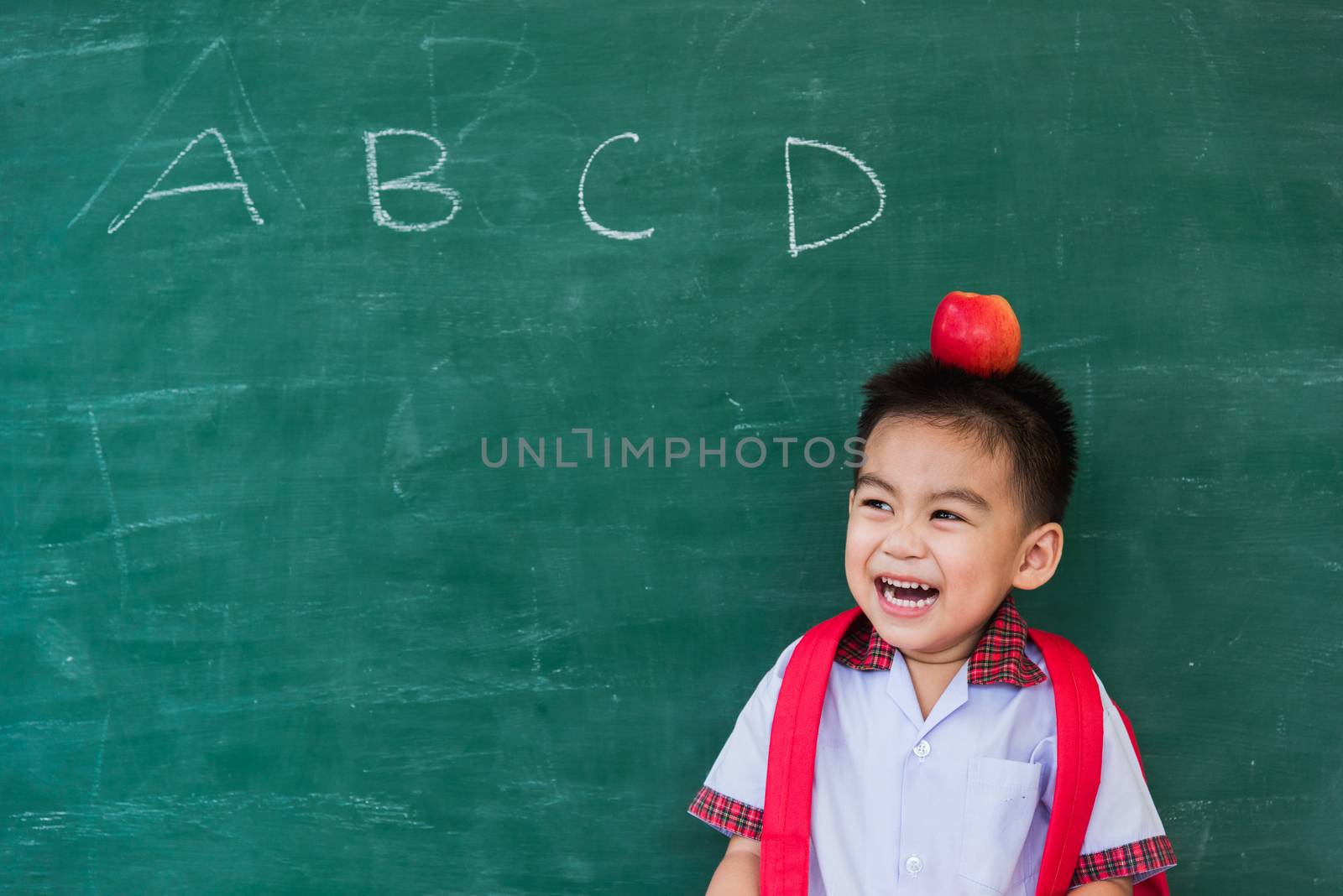 Back to School. Happy Asian funny cute little child boy from kindergarten in student uniform with school bag and red apple on head smiling on green school blackboard, First time to school education
