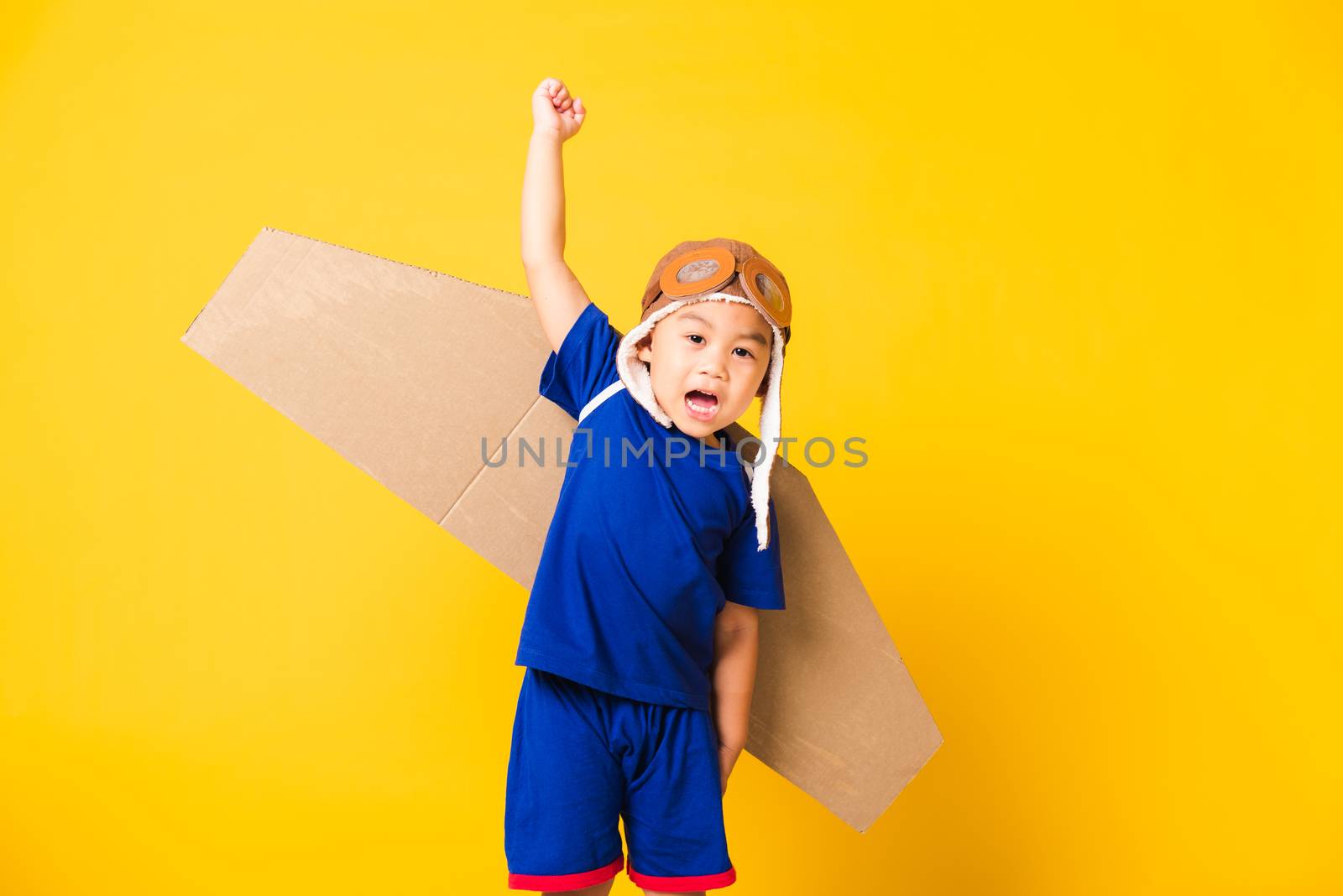 Happy Asian handsome funny child or kid little boy smile wear pilot hat play and goggles raise hand up with toy cardboard airplane wings flying, studio shot isolated yellow background, Startup freedom