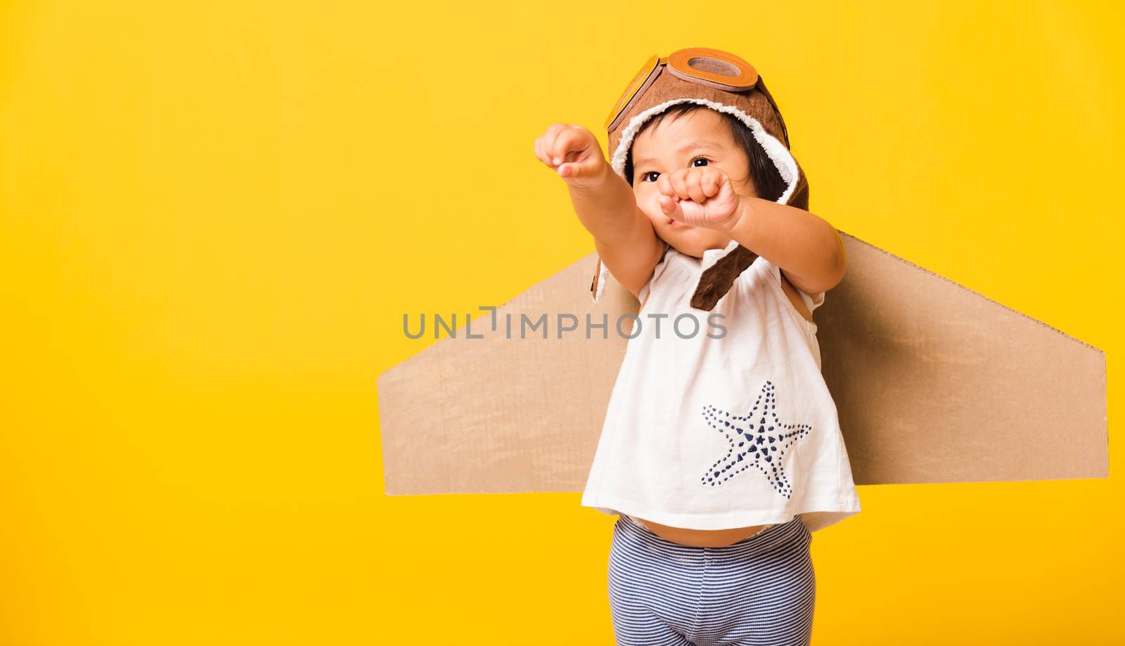 Happy Asian beautiful funny baby little girl smile wear pilot hat raise hand up play and goggles with toy cardboard airplane wings fly, studio shot isolated yellow background, Startup freedom concept