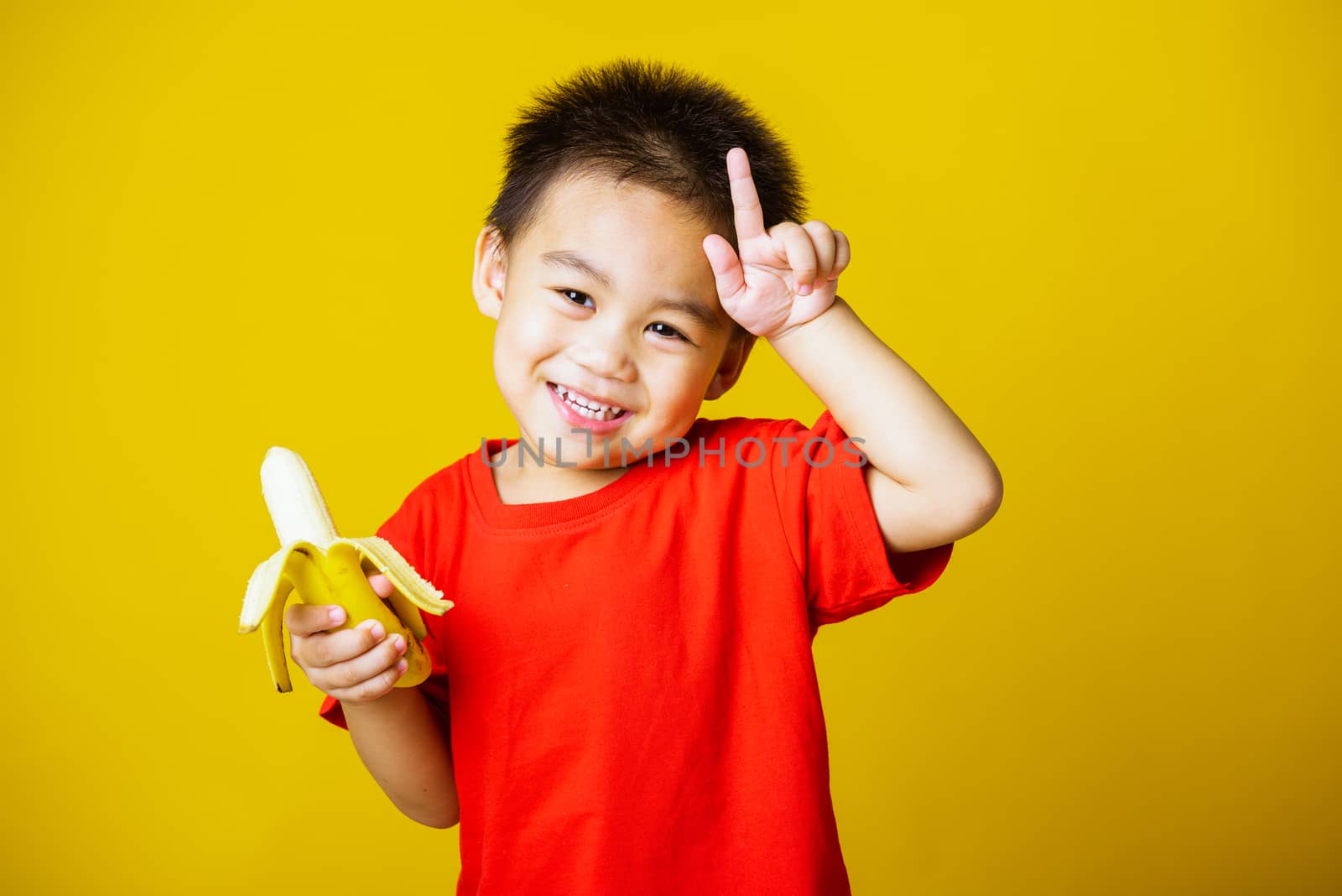 Happy portrait Asian child or kid cute little boy attractive smile wearing red t-shirt playing holds peeled banana for eating, studio shot isolated on yellow background