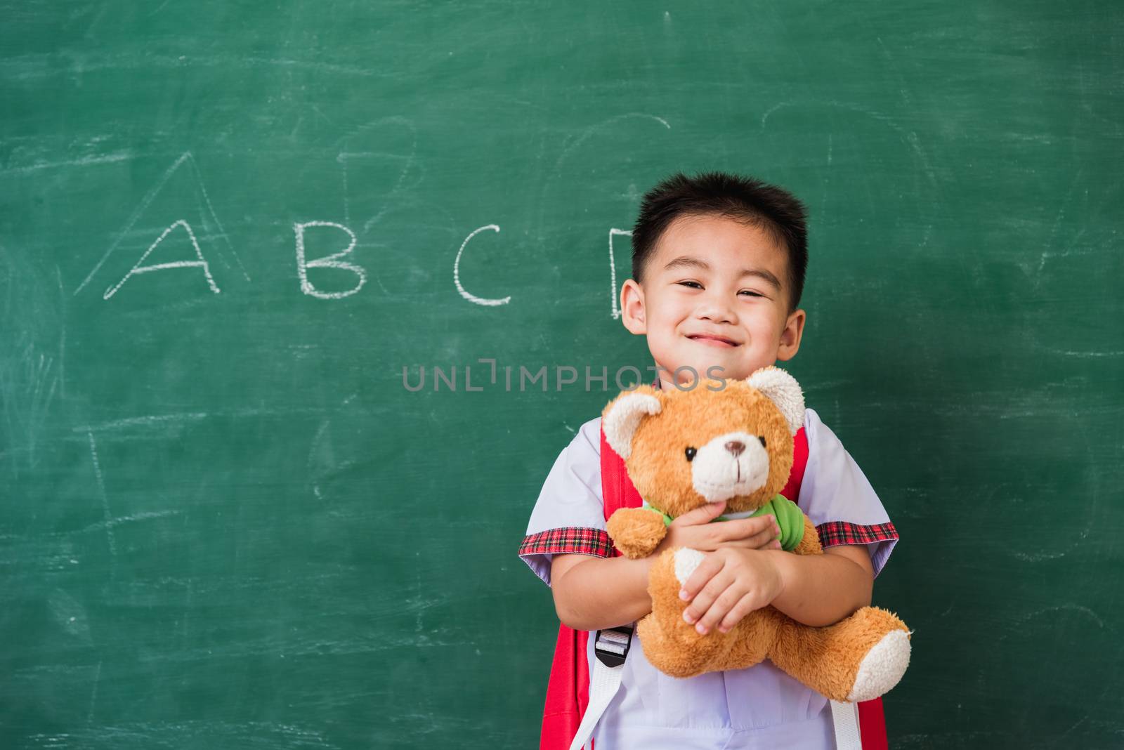 Back to School. Happy Asian funny cute little child boy from kindergarten in student uniform with school bag smiling and hugging teddy bear on green school blackboard, First time to school education