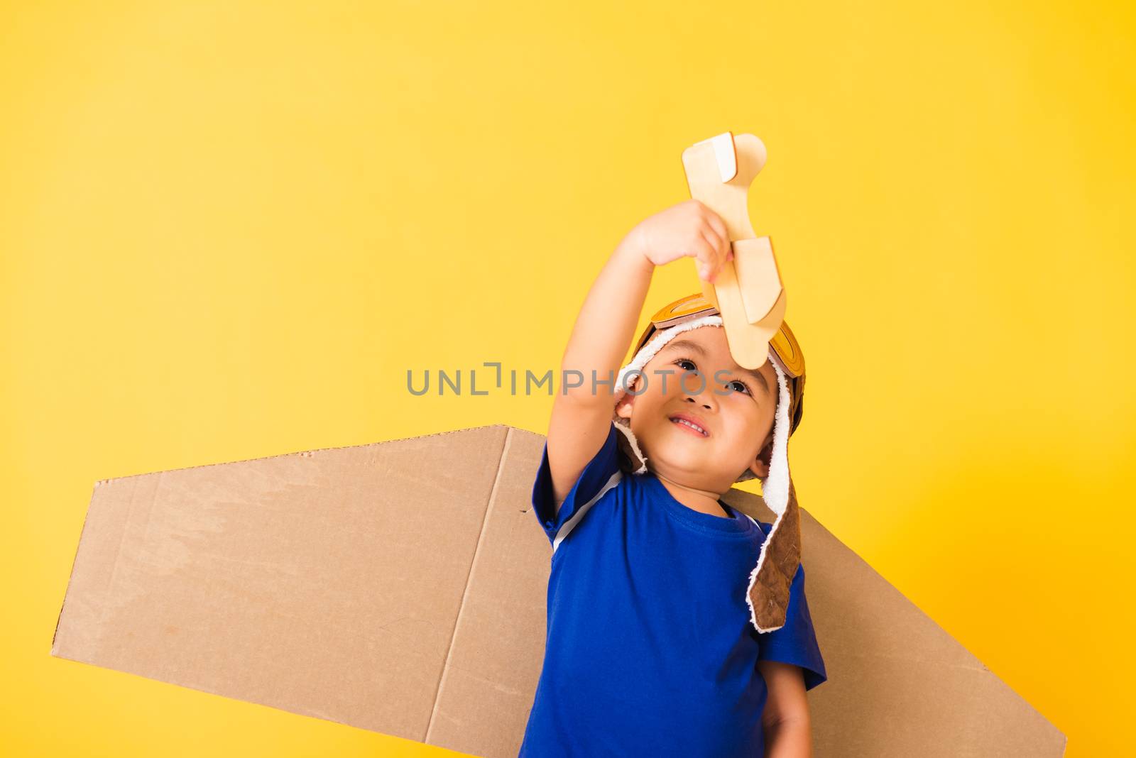 Happy Asian handsome funny child or kid little boy smile wear pilot hat play and goggles with toy cardboard airplane wings fly hold plane toy, studio shot isolated yellow background, Startup freedom