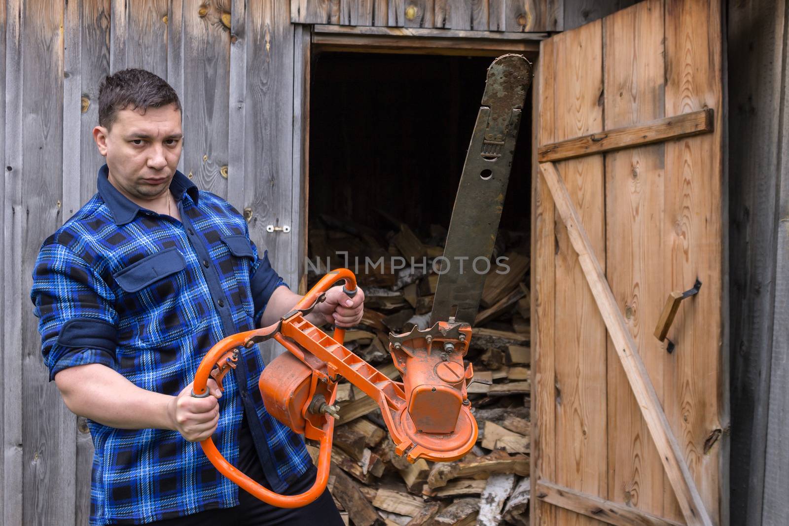 Farmer in a blue shirt with a big chainsaw on a background of a wooden warehouse