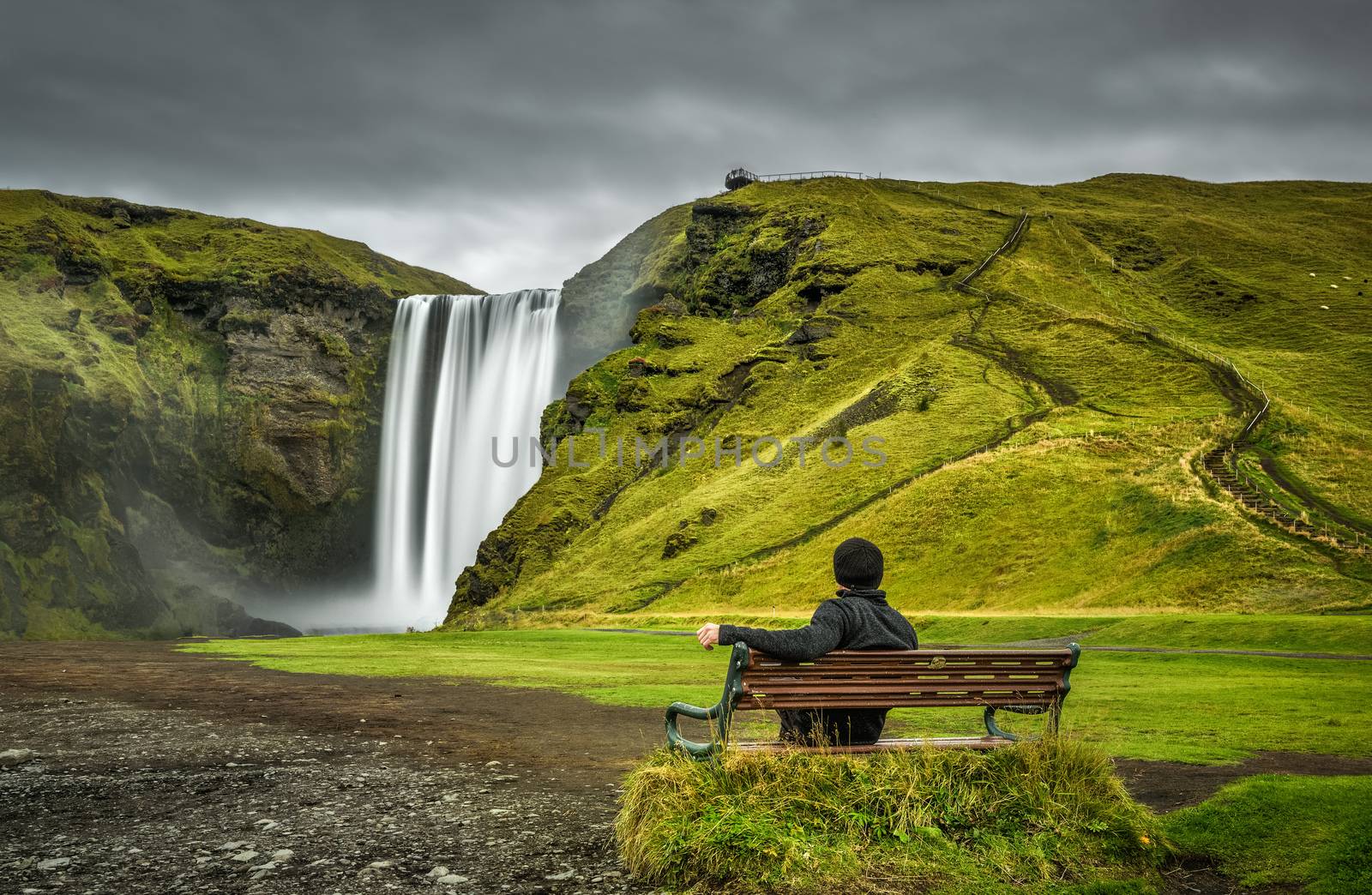 Hiker sits on a bench and looks at the famous Skogafoss waterfall in southern Iceland