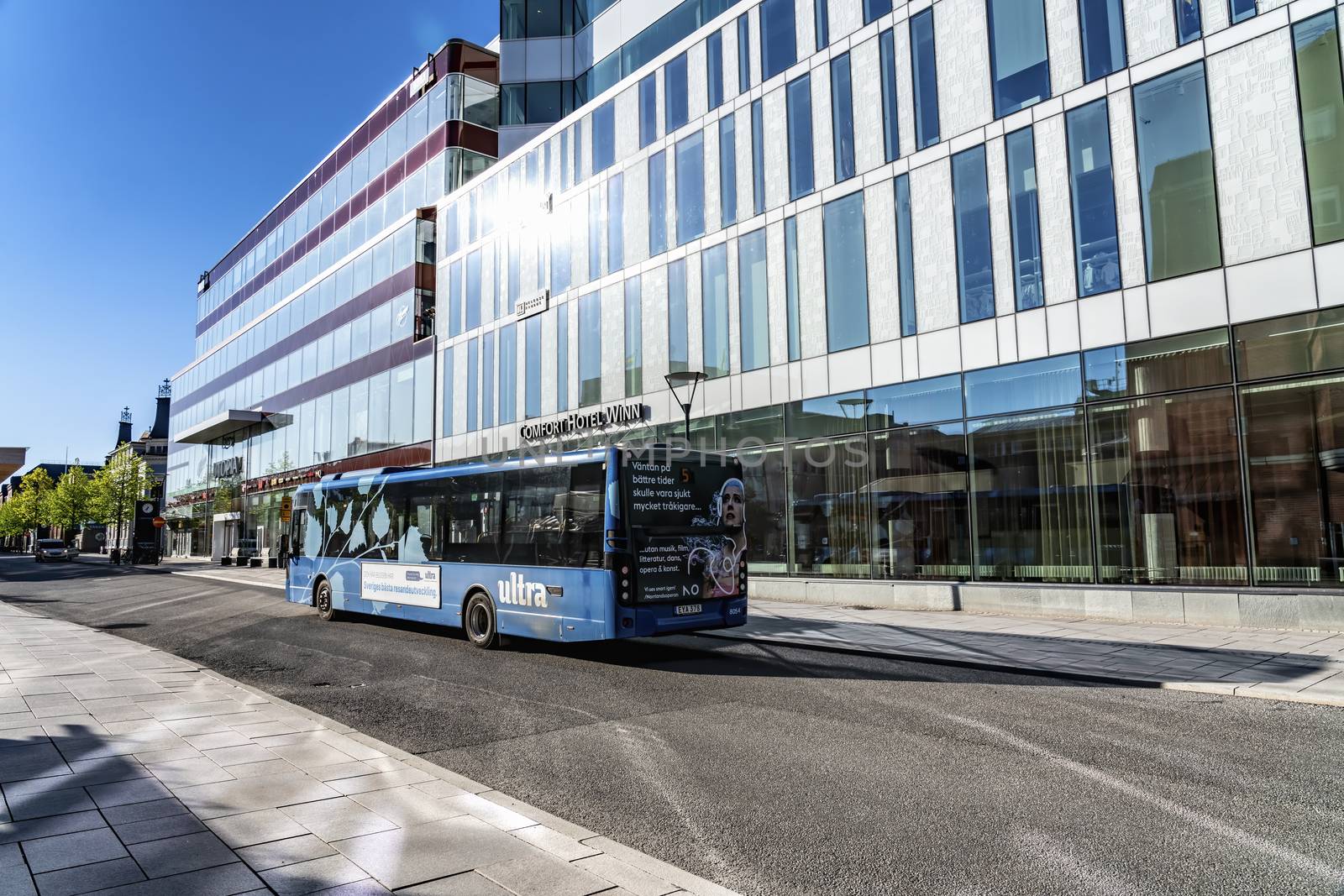 UMEA, SWEDEN - JUNE 10, 2020: Outdoor view of blue bus drive close to high modern hotel and shopping centrum building, Umea downtown. Early summer sun reflects in windows, morning. Umea, Vasterbotten by skydreamliner