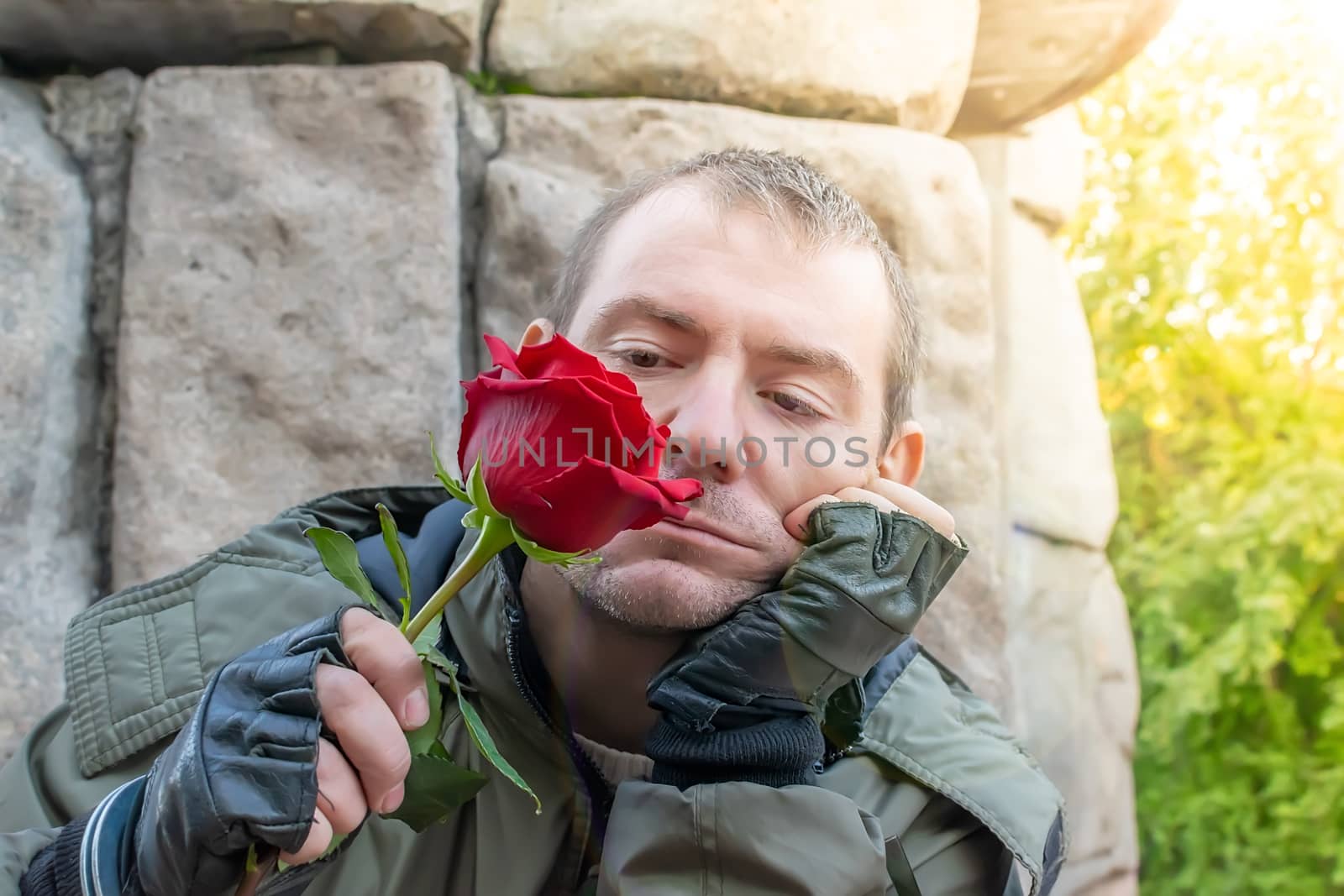 a guy in a jacket and leather gloves sits, sad and sniffs a red rose bud in his hand
