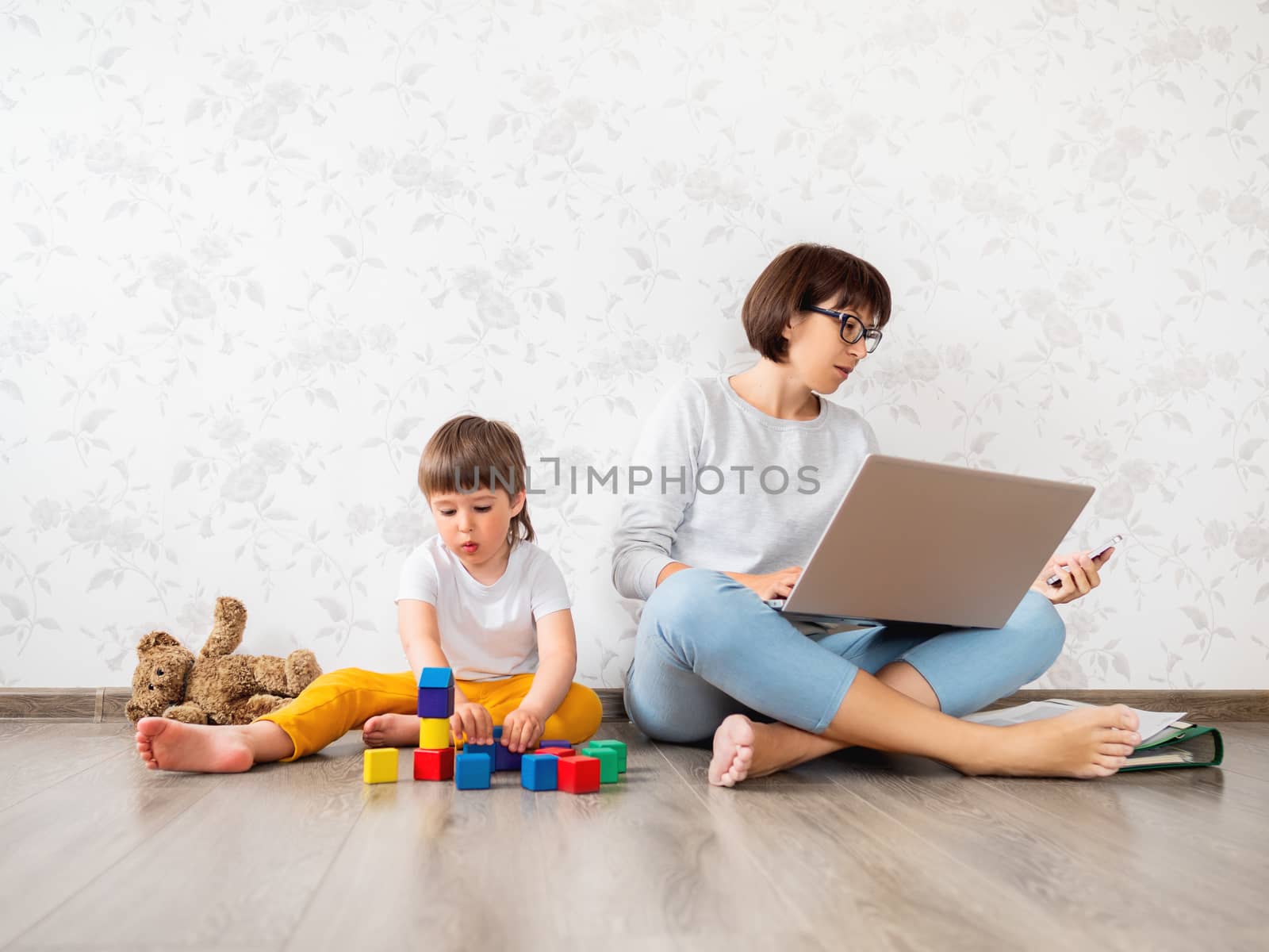 Mom and son at home quarantine because of coronavirus COVID19. Mother works remotely with laptop and smartphone, son plays with toy blocks. Self isolation at home.