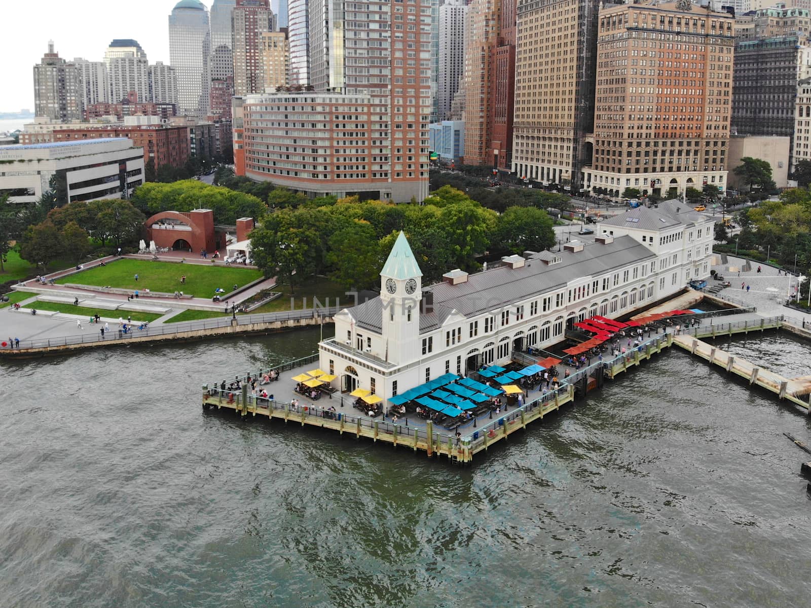 Aerial view of Battery Park pier A leading to Liberty Island, people can be seen waiting in line for boarding as well as looking at Liberty Island located opposite to the docks. October, 22nd, 2019