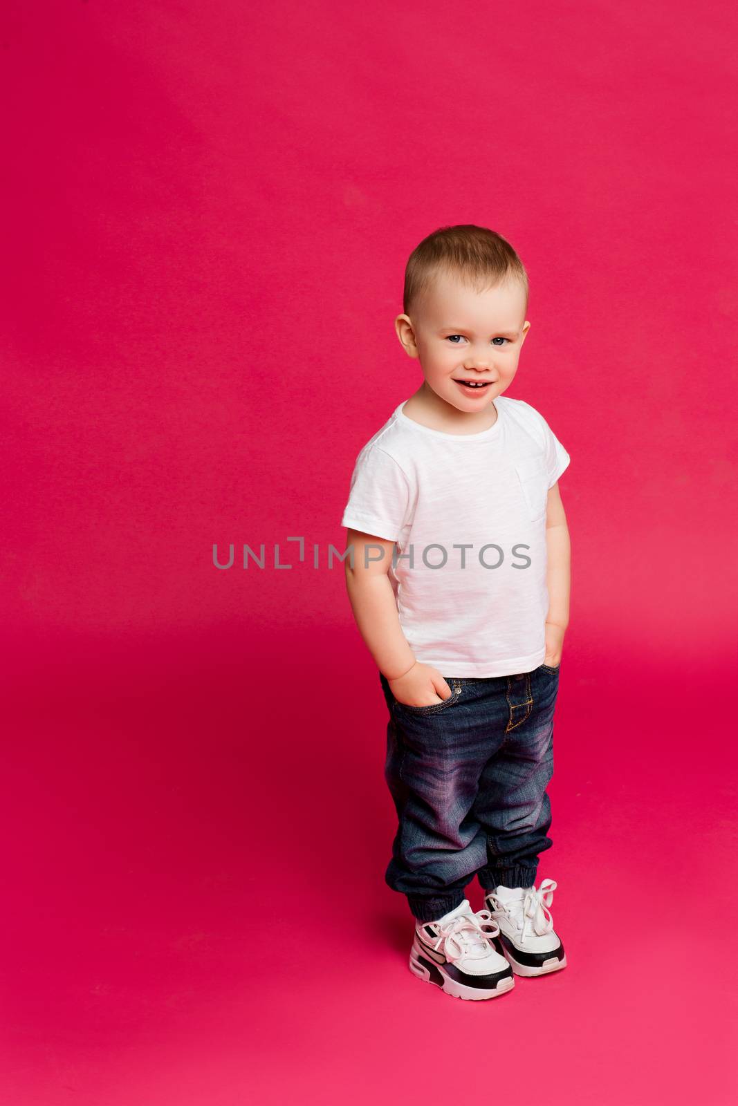 Portrait of stylish little blond boy wearing in jeans and white t shirt posing at camera, holding arms in pockets and smiling. Fashionable kid at pink studio.