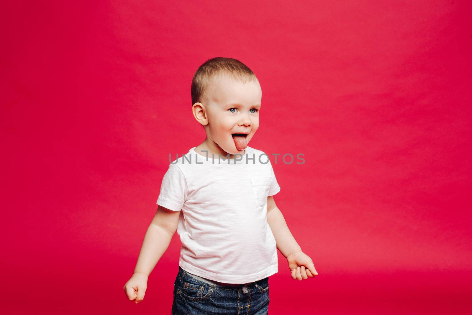 Sweet little emotion boy playing at studio and showing tongue out, looking away. Kid stick out and making face, posing away. Pink studio background. Concept kids fashion.