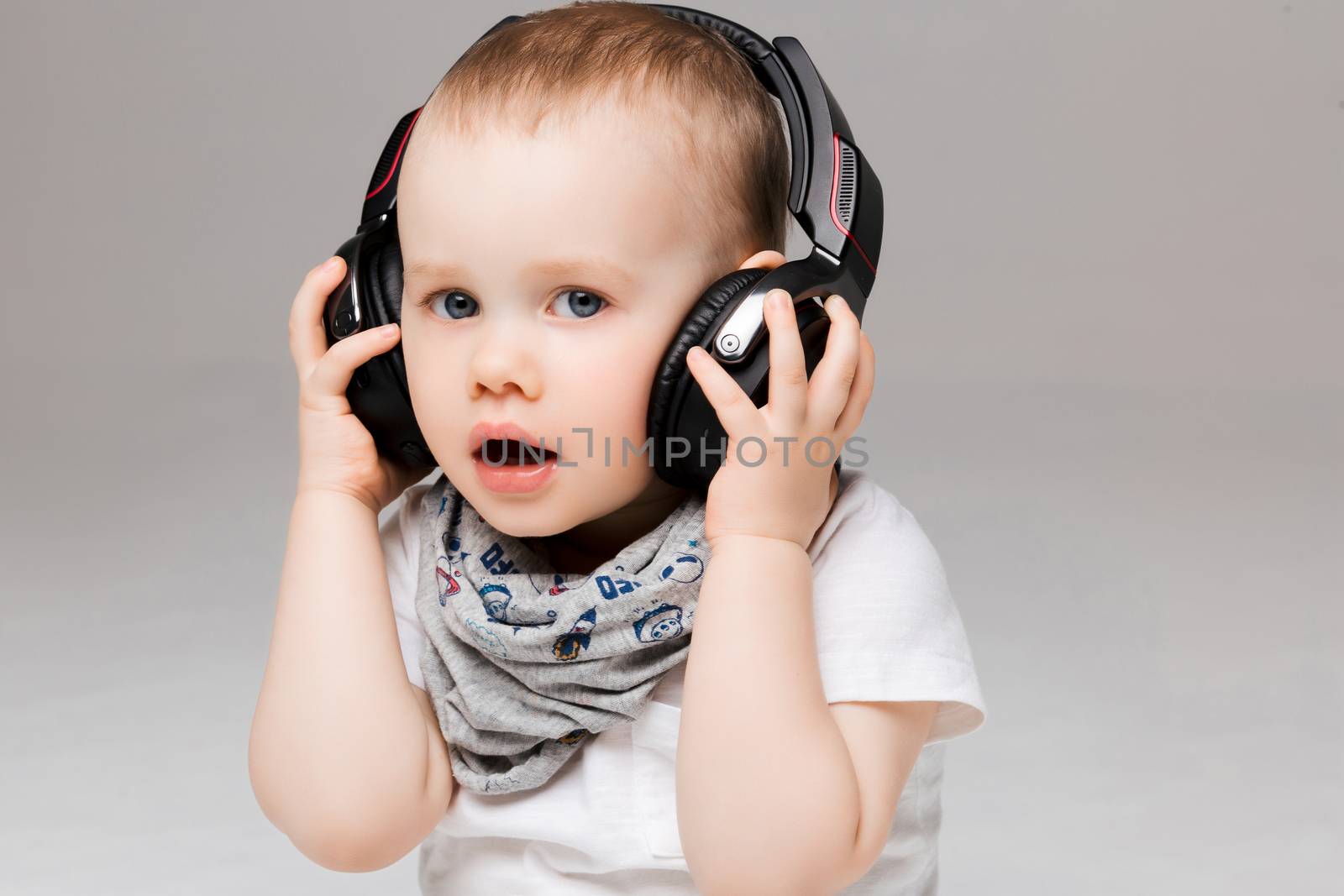 Studio portrait of little surprised boy listening music in big earphones with opened mouth, interesting looking at camera. Stylish sweet kid stylish wearing, posing at gray studio.