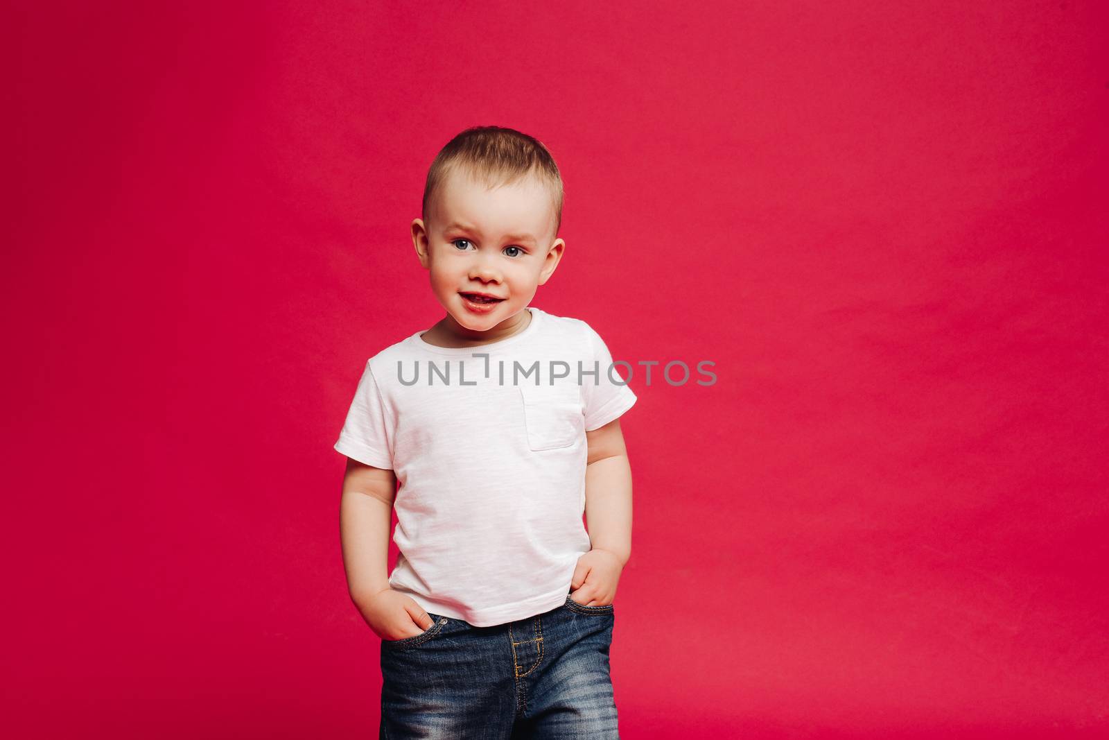 Emotionaly stylish little boy wearing in jeans and white t shirt holding hands in pockets and smiling. Fashionable child two year old posing at studio against pink background.