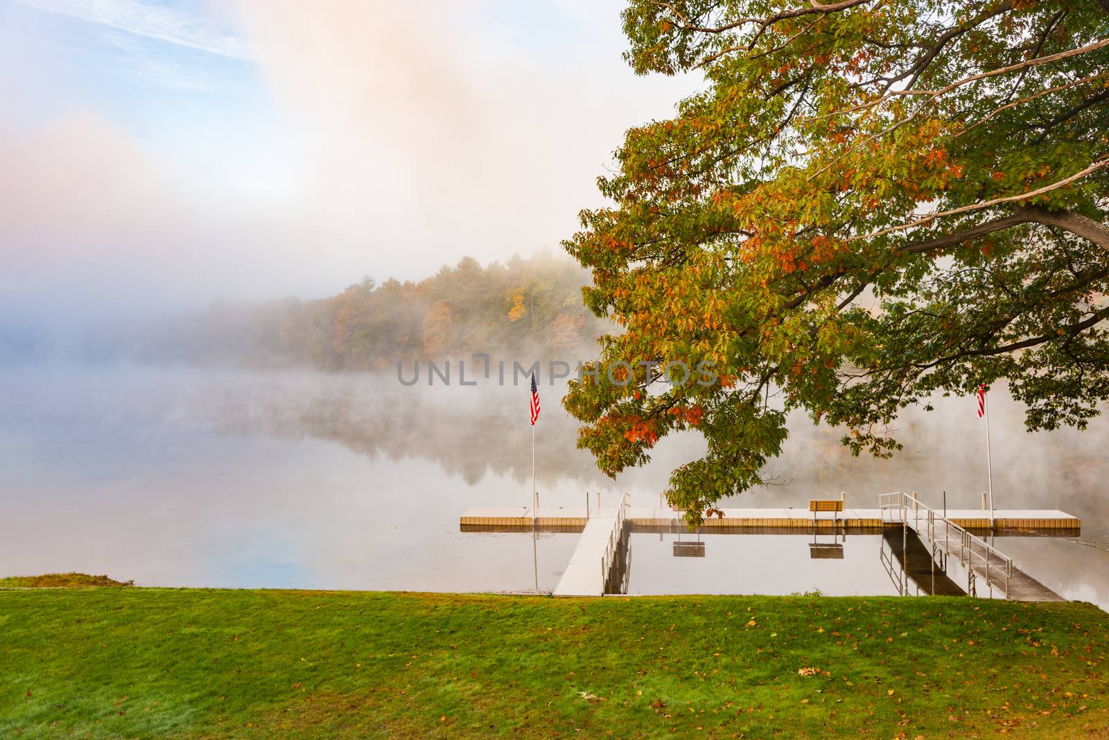 Scenic river edge and T-shaped pier under oak tree early morning fog just above the water level of Connecticut River with colorful fall foliage along both sides converging into distance.