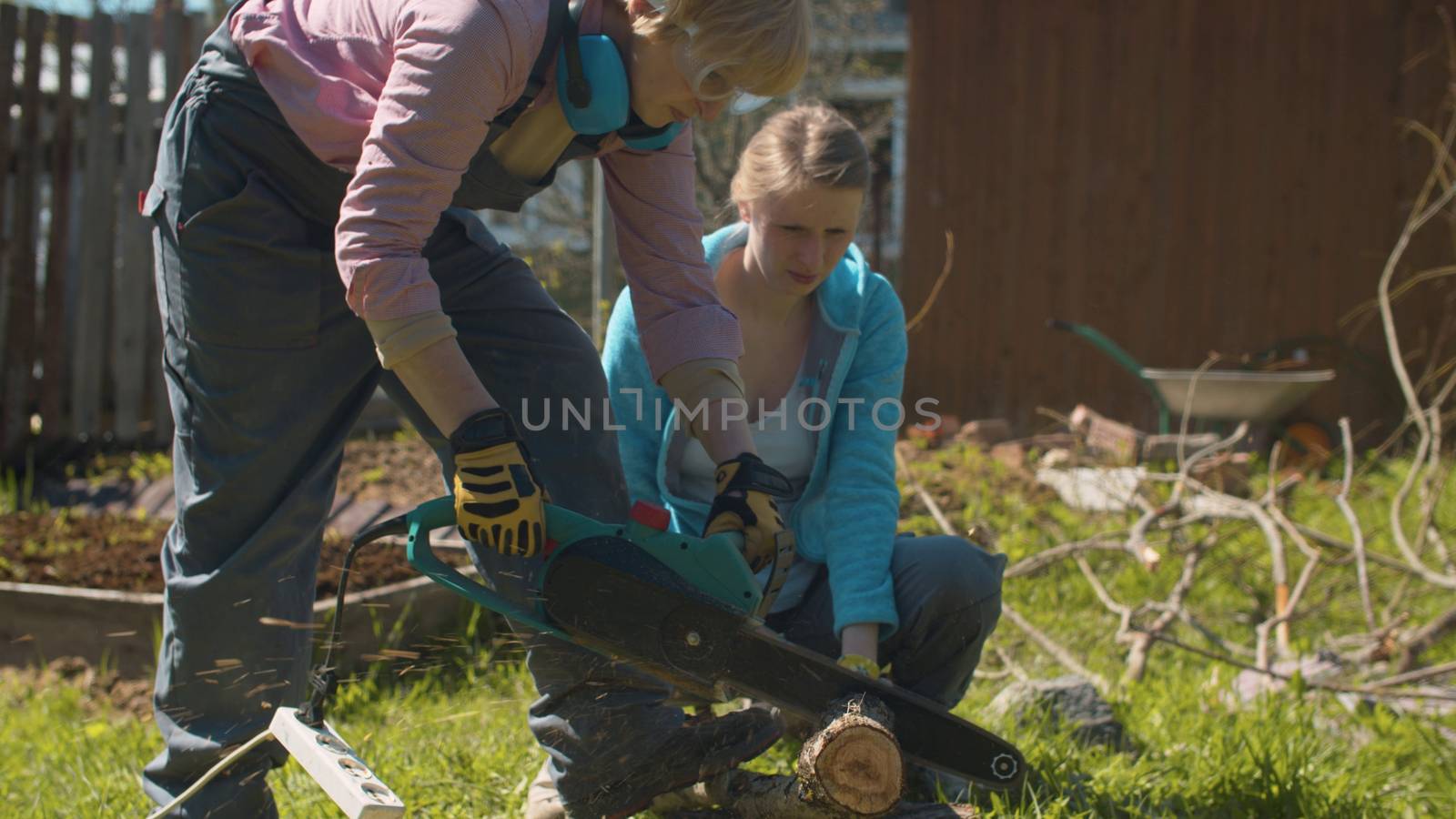 Two women working in the courtyard of country house. Mature cutting tree with an electric chain saw, young helping she. Active and healthy lifestyle concept