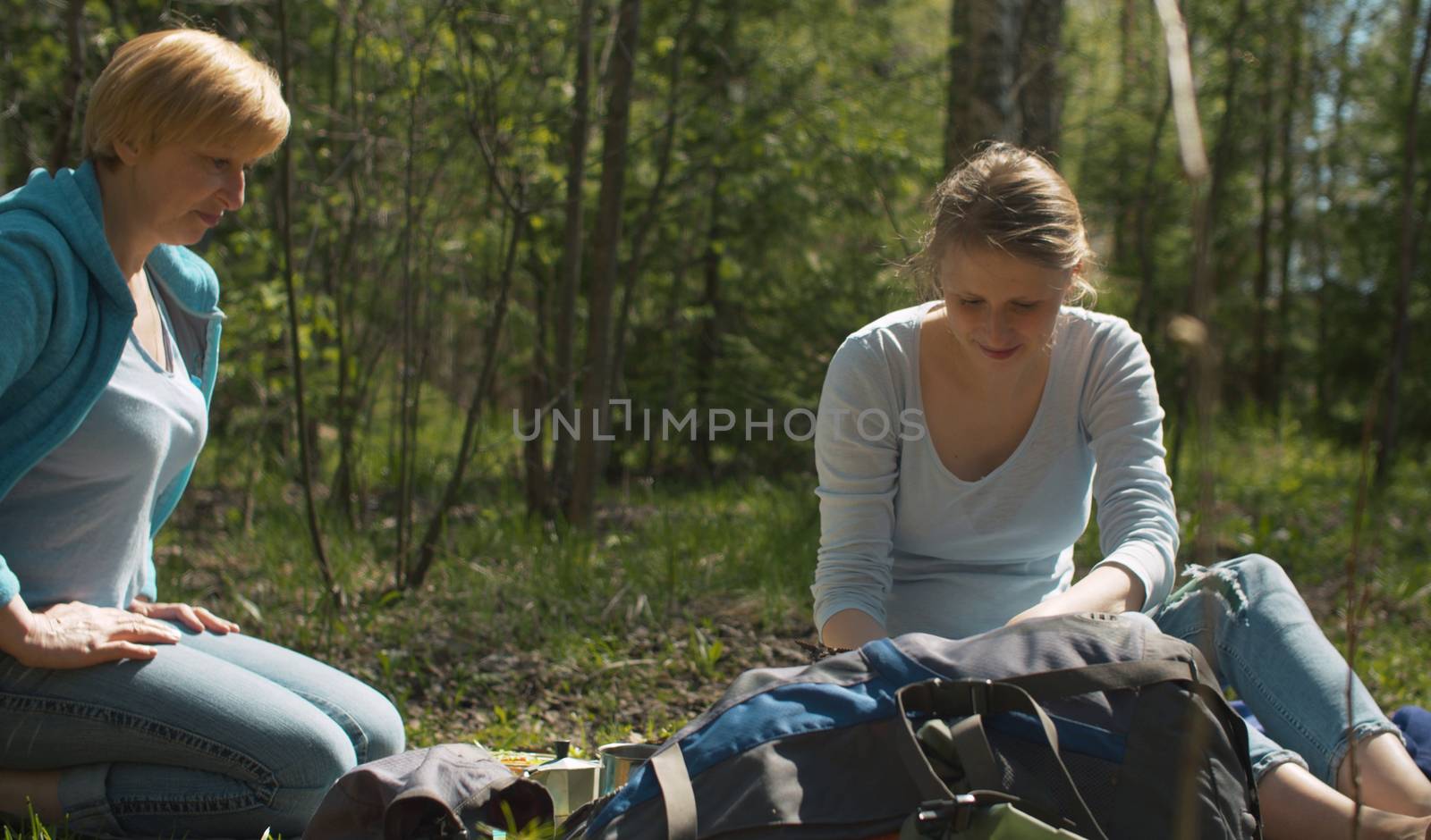 Snack during trekking. Two women sitting on the grass in the forest. They take out a italian espresso maker and a lunch box from a backpack.