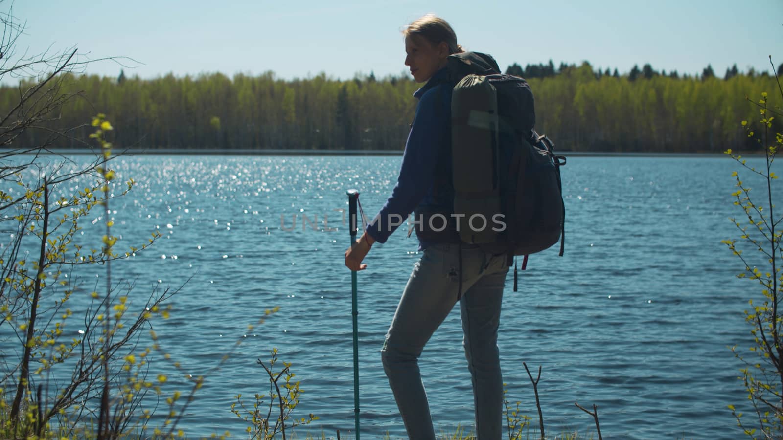 Young woman with a backpack on the shore of a forest lake. Beautiful glare from the sun on a water ripple. Trekking in the forest, active and healthy lifestyle concept