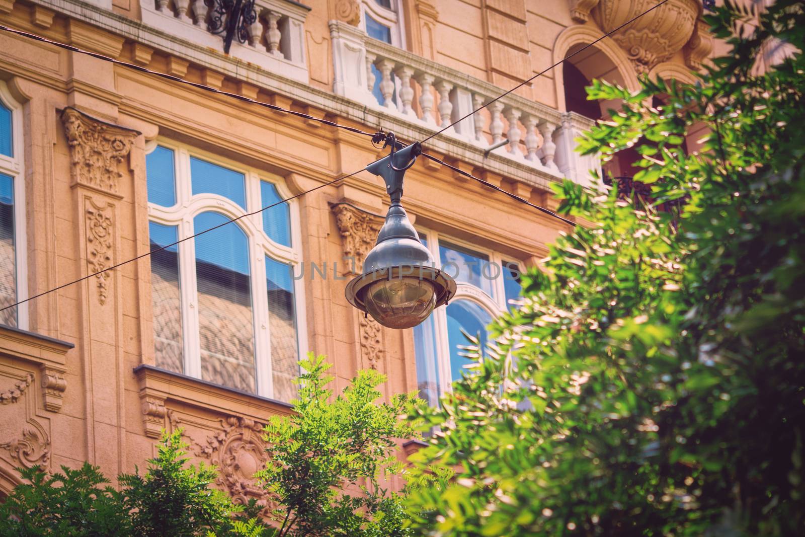Close-up of a street lamp on aerial wires above green trees and a beautiful old building in the background. Hanging street light in Budapest city center.