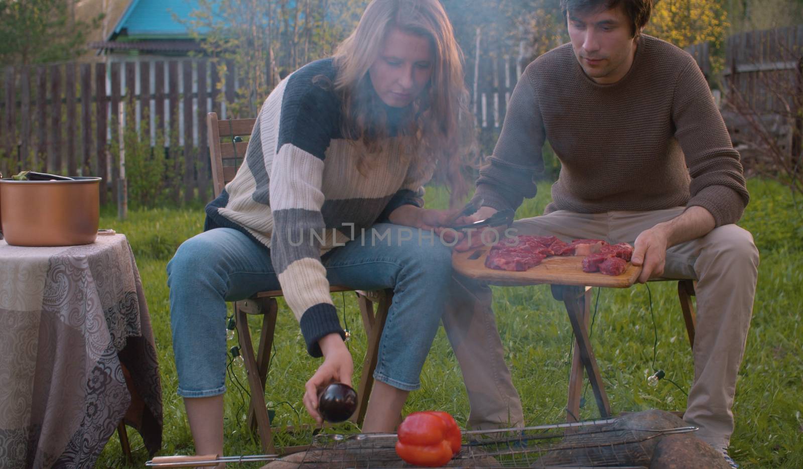 Couple cooking on barbecue grill at spring outdoor. Woman putting vegetables on the grill in the courtyard of a country house. Leisure, food, people and holidays concept