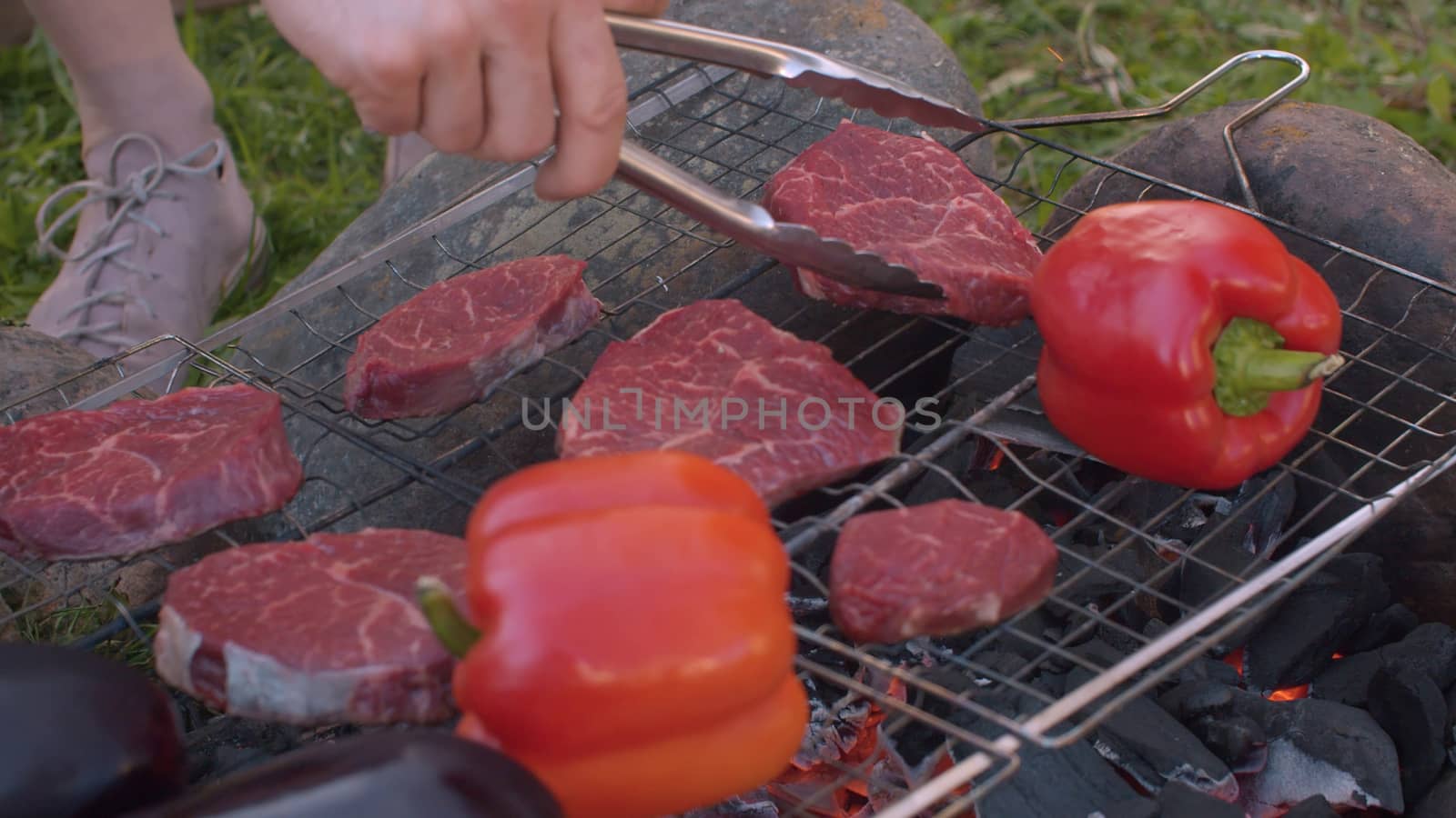 Close up hand putting piece of meat on the grill. Cooking of beef and vegetables on barbecue grill outdoor. Leisure, food, people and holidays concept. Top view