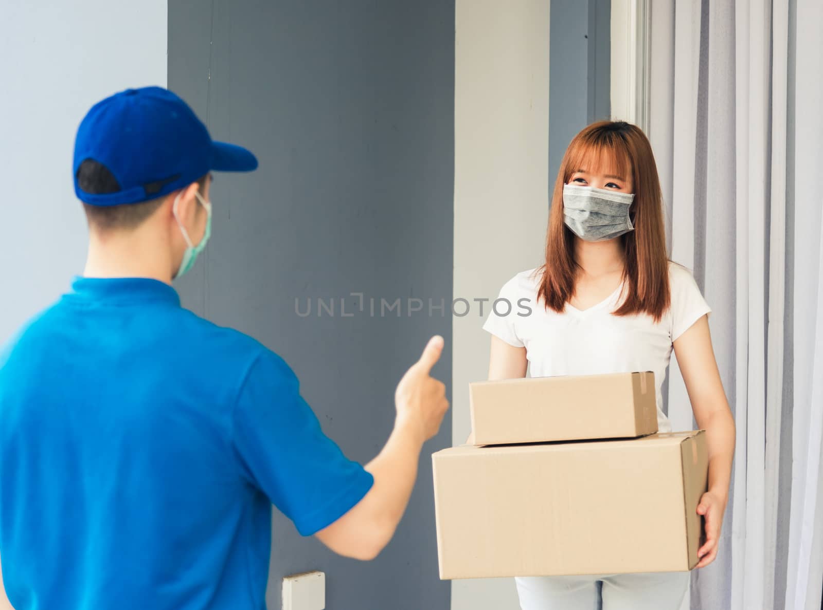 Asian delivery express courier young man giving parcel boxes to woman customer receiving both protective face mask and show thumbs up finger for good support sign, under curfew pandemic coronavirus