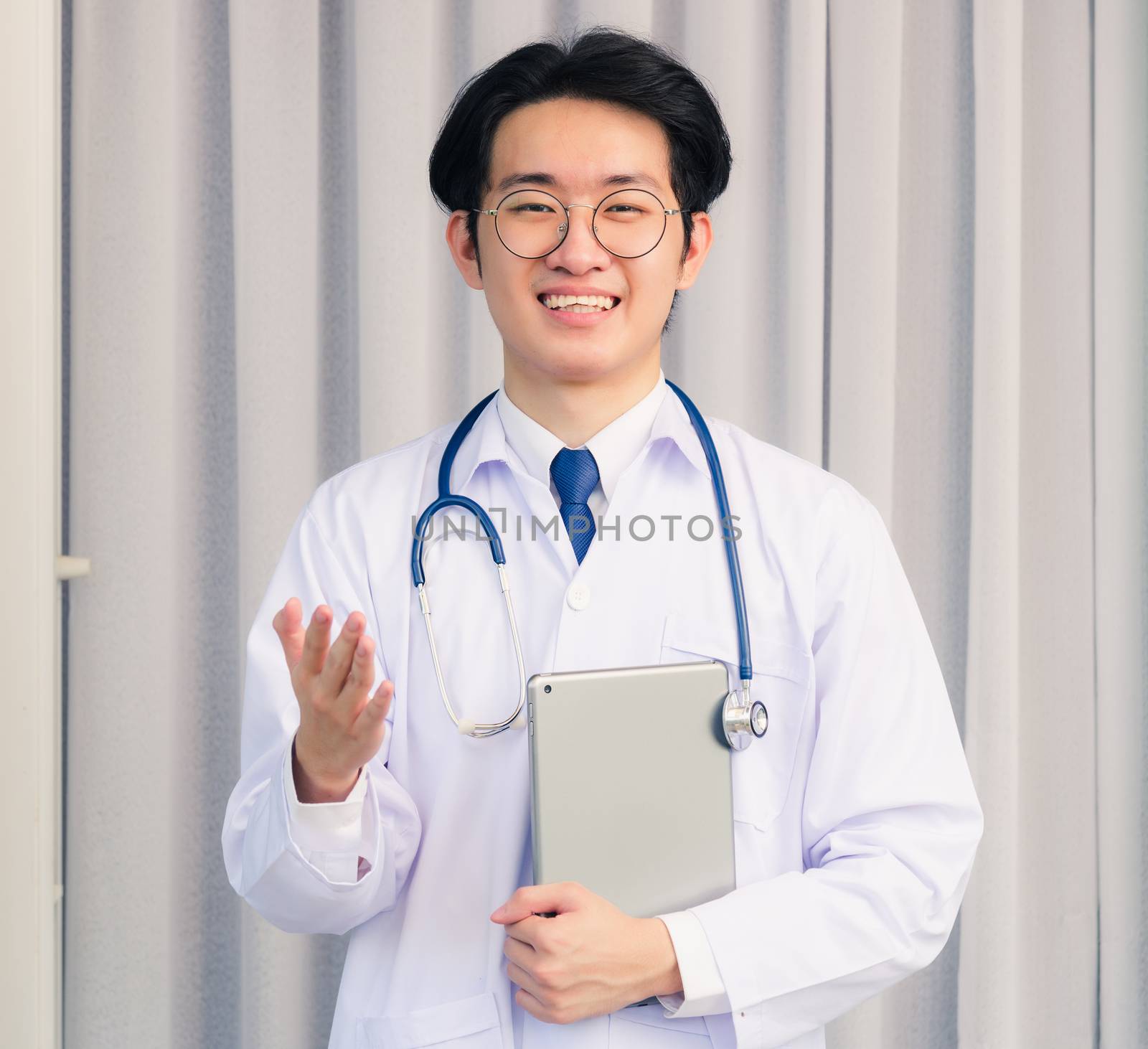 Portrait closeup of Happy Asian young doctor handsome man smile in uniform and stethoscope neck strap holding smart digital tablet on hand and show hand presence to front, healthcare medicine concept