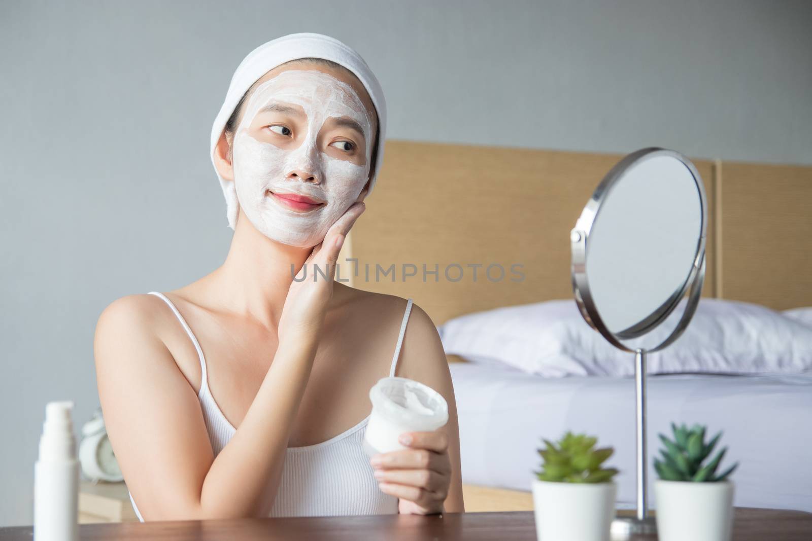 Asian woman applying moisturizer face scrub peeling white clay mask on skin, looking in mirror with smiley face using cream, touching face. self skin care beauty treatment at home by asiandelight