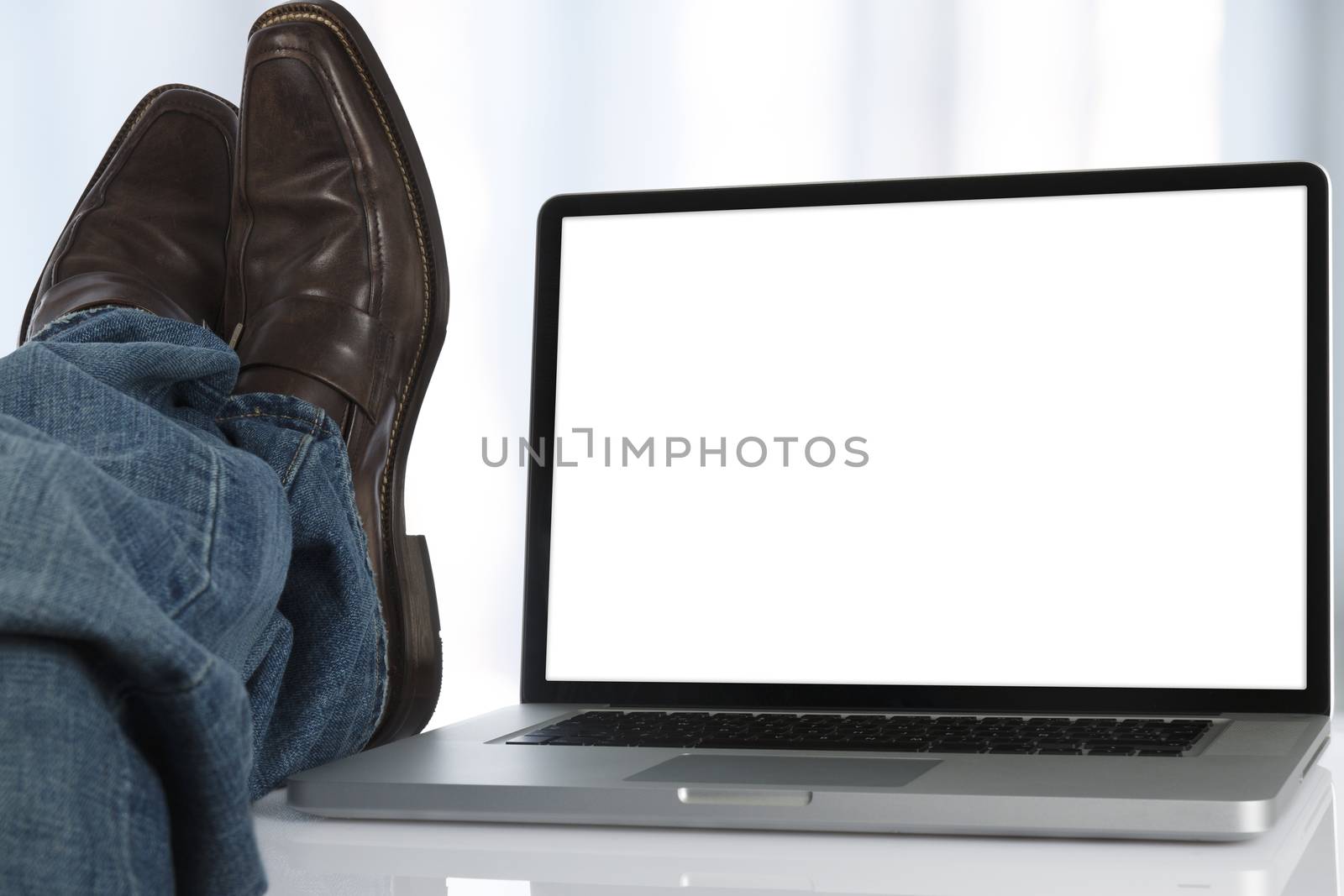Man sits with his feet on the table next to a laptop with white screen.