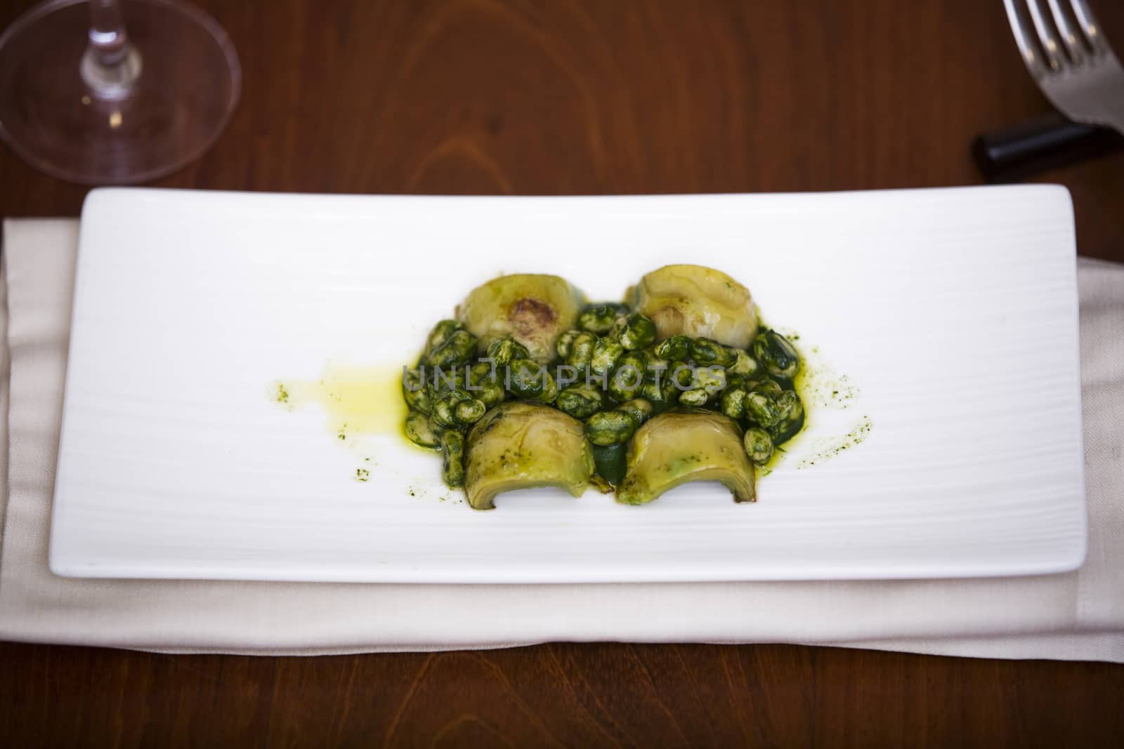 Delicious dish of broad beans (fava beans) and artichokes dressed with spinachs and olive oil. The main color is green and the plate is white over a background of wood. There is a fork and a glass.