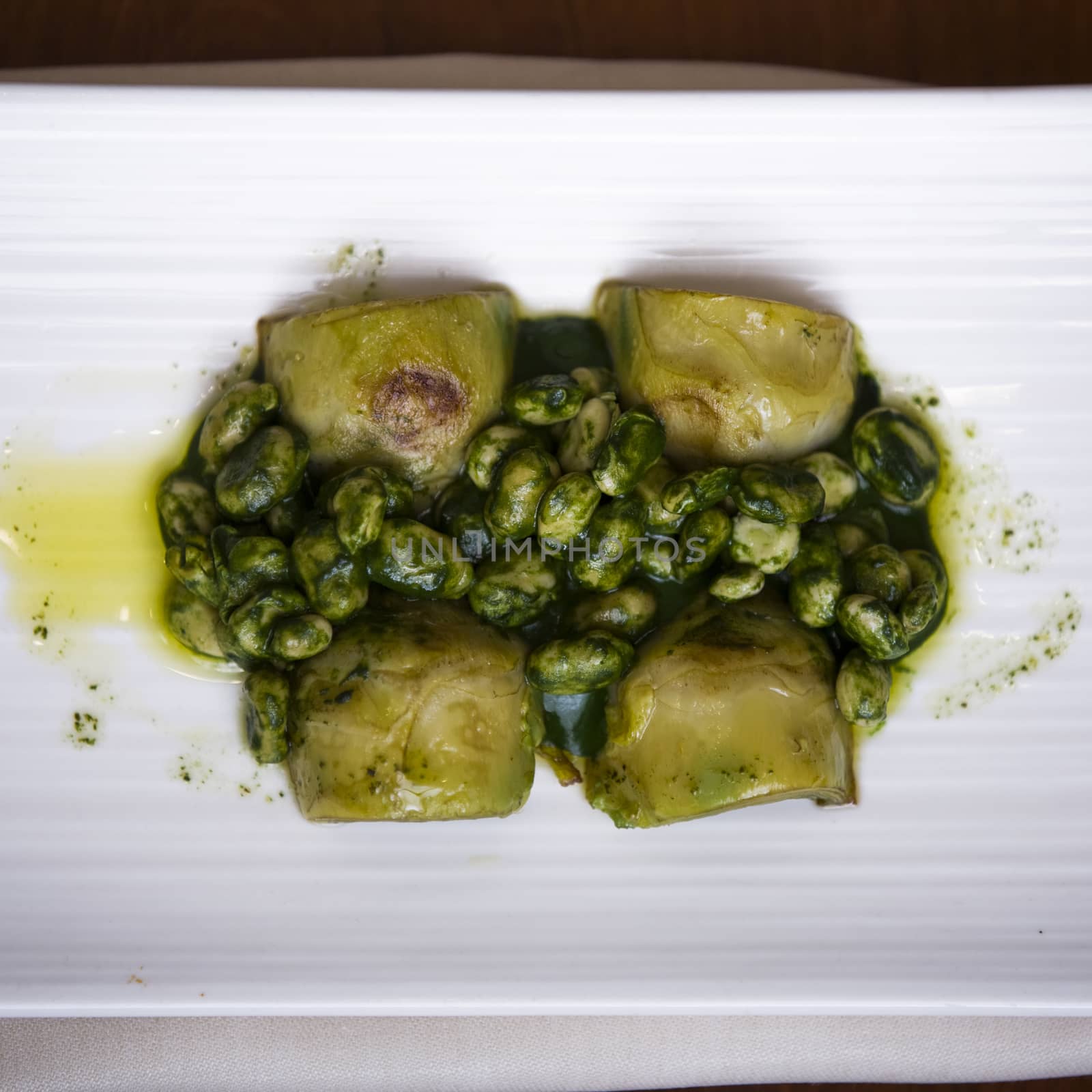 Delicious dish of broad beans (fava beans) and artichokes dressed with spinachs and olive oil. The view is an overhead shot, the main color is green and the plate is white over a background of wood.