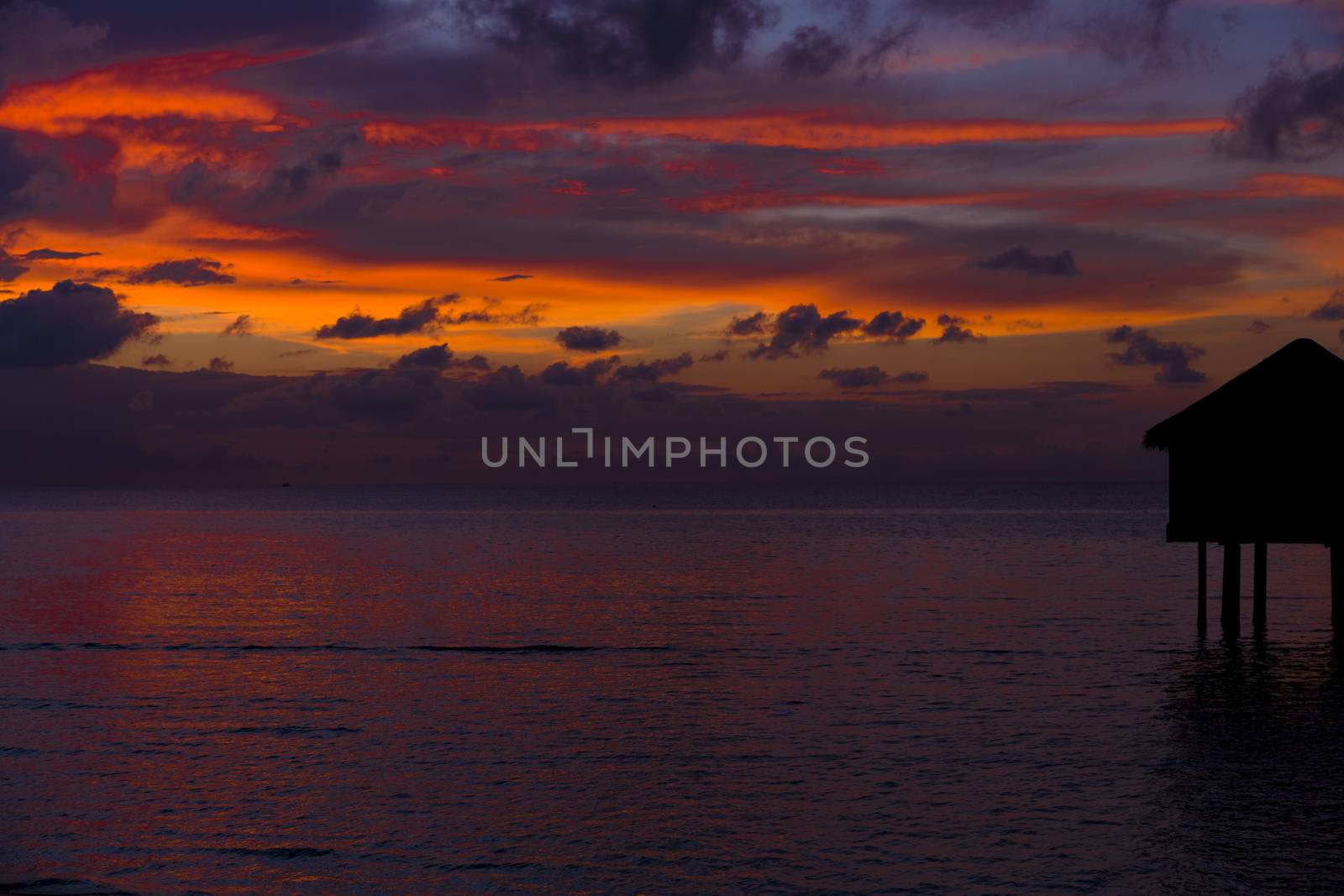 Silhouette of a bungalow over the sea in front of the sunset in the Maldives. The bungalow is situated in the right of the frame. There is an amazing contrasts of tones with the colors and the clouds.