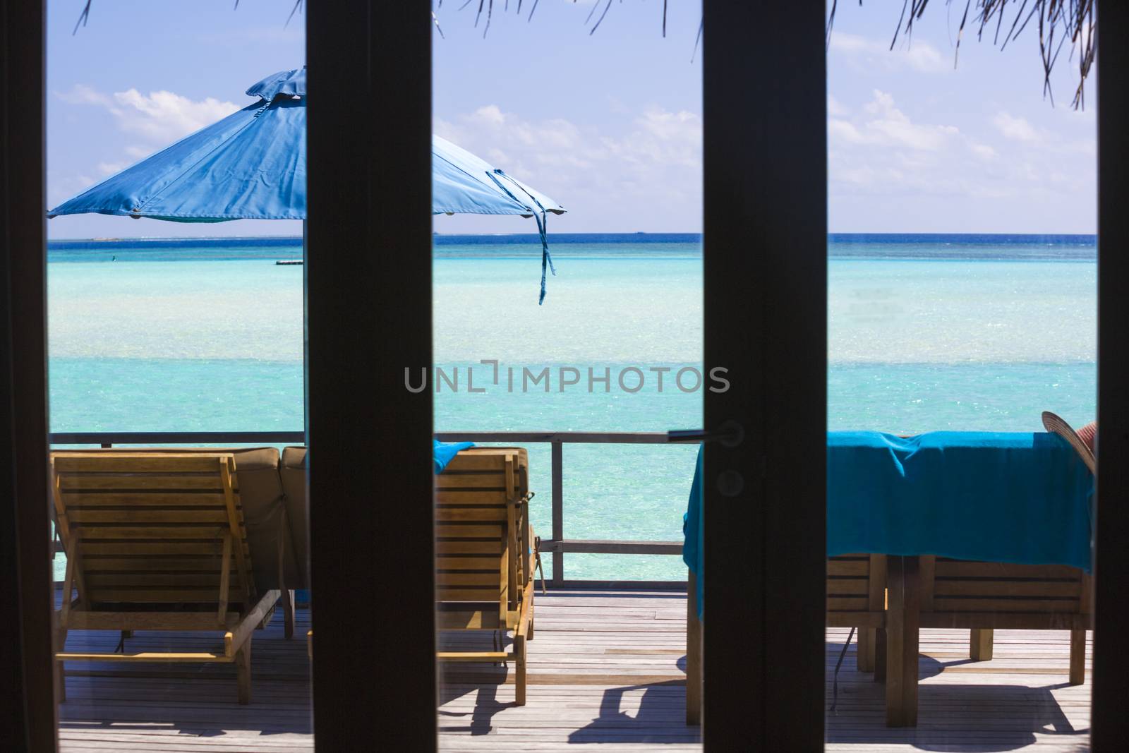 View from the inside of the bungalow through the window  in The Maldives. On the Deck there are deck chairs and beach umbrella. It is a beuatiful view of the sea, the water is clear, turquoise and in a strong contrast depending on the depth.