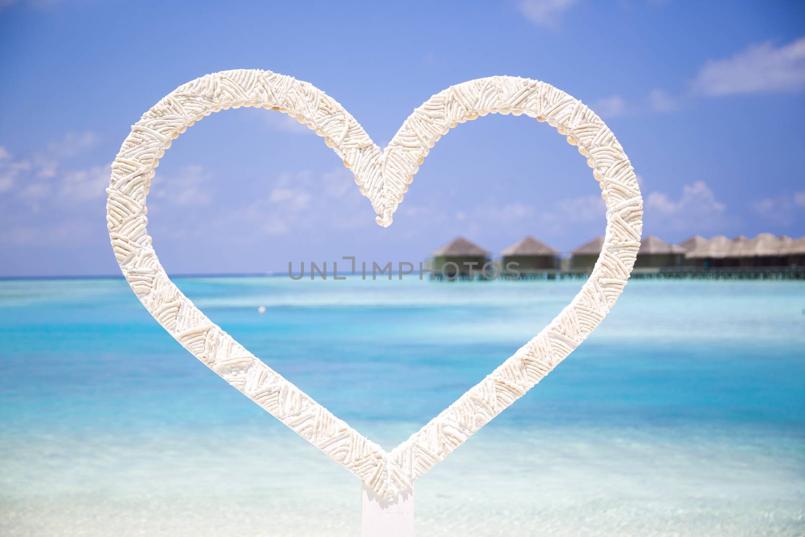 Heart to shoot photos of honeymoon couples in the beach in Maldives with a tropical background with bungalows over water. It is a beautiful sunny day with few clouds.
