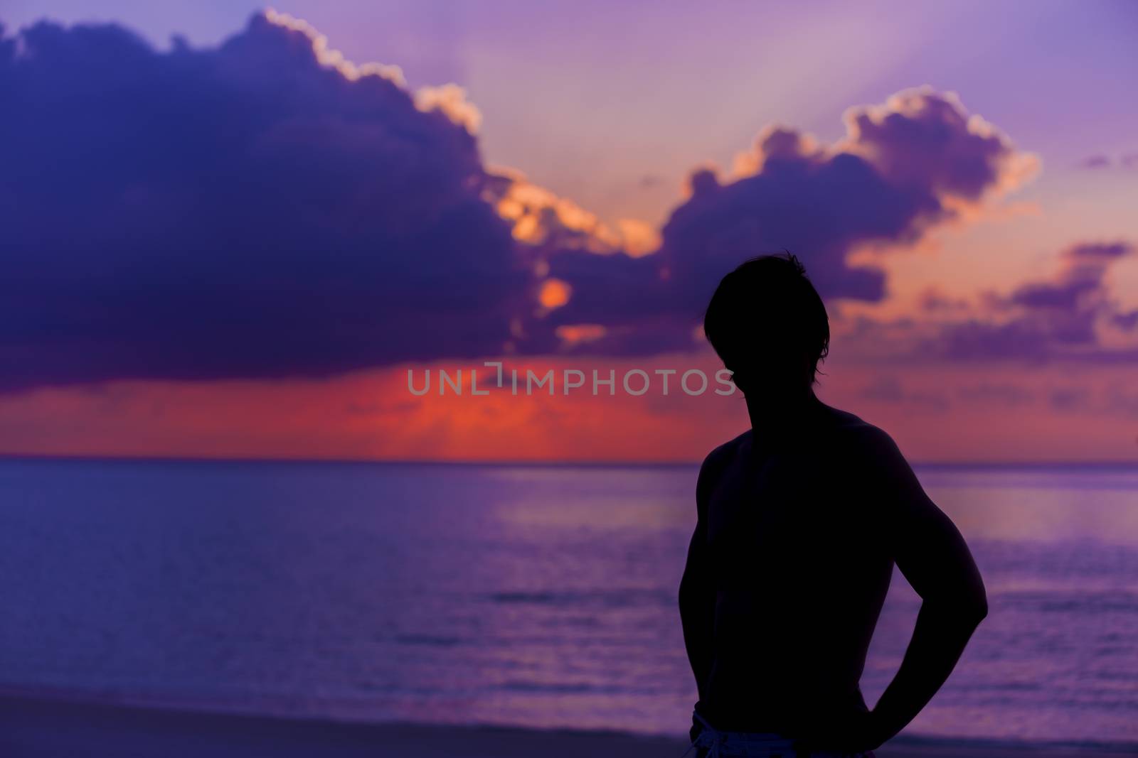 Beautiful idyllic seascape in The Maldives at sunset. A silhouette of the man is standing in front of the sun when it is getting darker and darker. The colors of the image are purple, blue and orange in a perfect contrast with the man in the beach.