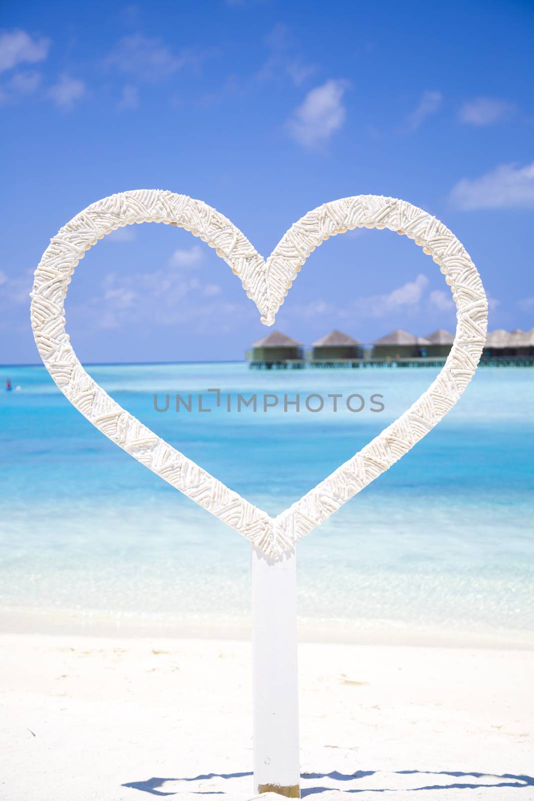 Heart to shoot photos of honeymoon couples in the beach in Maldives with a tropical background with bungalows over water. It is a beautiful sunny day with few clouds.